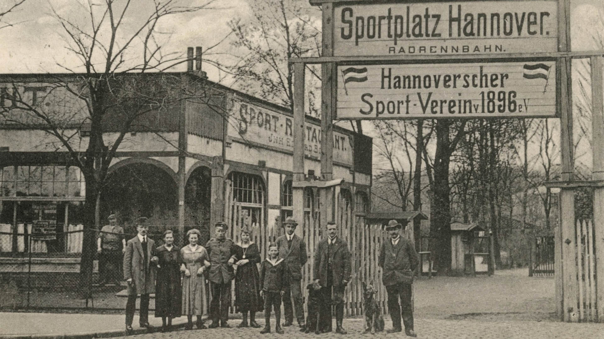 Schwarz-Weiß-Foto von einer Gruppe von Menschen mit zwei Hunden vor einem Schild mit der Aufschrift "Sportplatz Hannover Radrennbahn - Hannoverscher Sportverein v 1896 eV ".