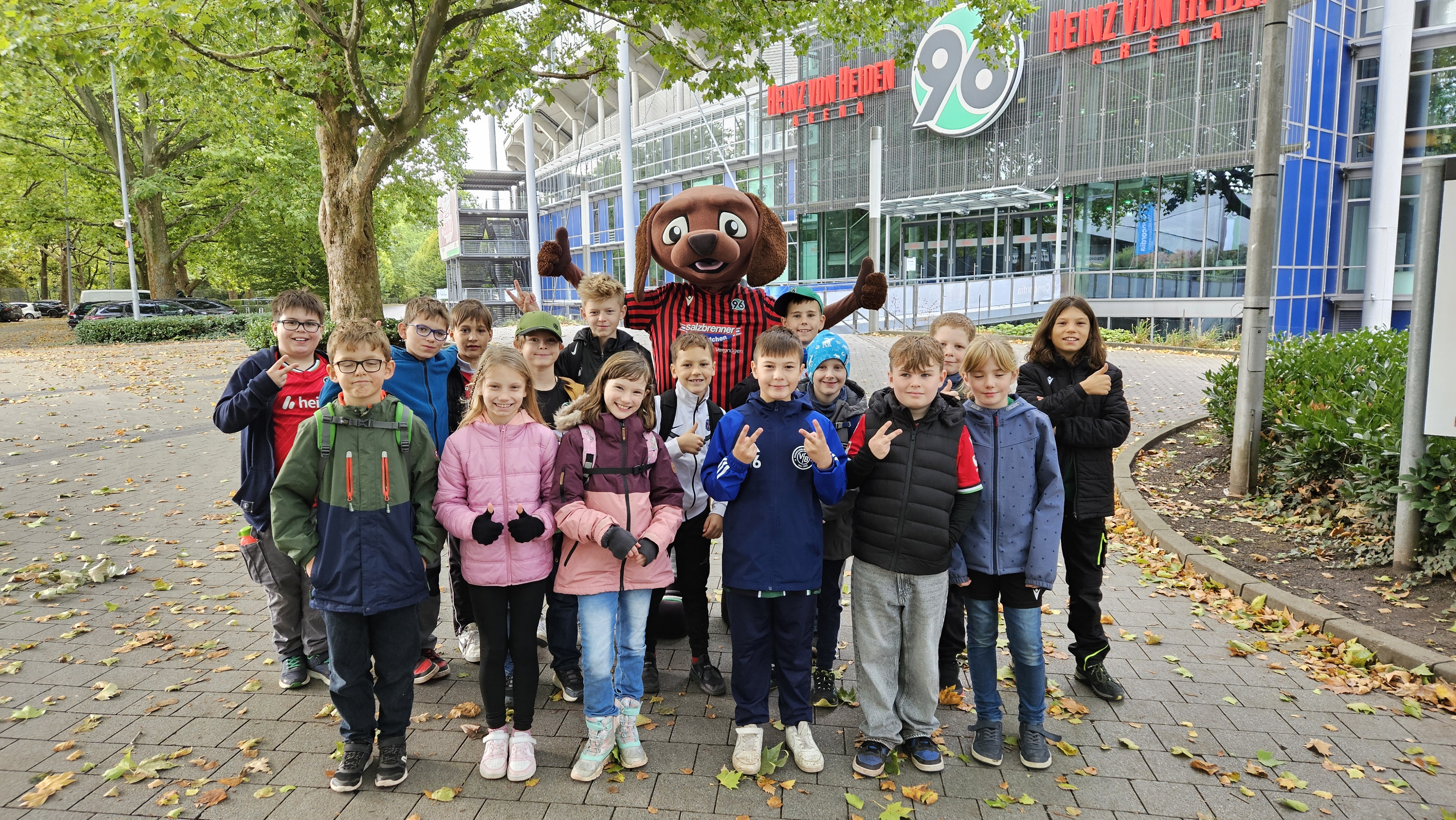 20 Kinder gucken lächelnd mit dem Maskottchen vor der Heinz von Heiden Arena.