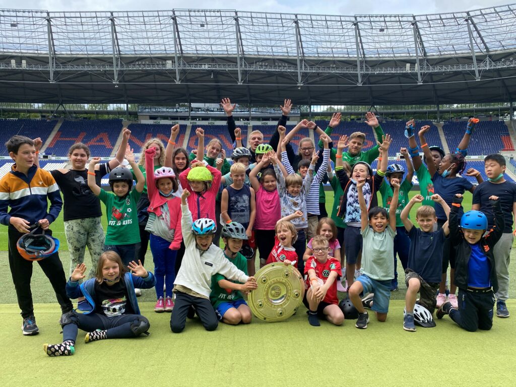Eine große Gruppe von Kinder stehen während des Ausflugs ins Stadion am Spielfeldrand und posieren für ein Foto gemeinsam mit der Meisterschale.