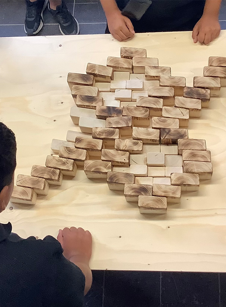 Mehrere Kinder stehen um einen hellen Tisch und legen kleine, rechteckige Holzklötze dicht nebeneinander. Die unterschiedlich gefärbten Holzstücke bilden das Wappen von Hannover 96.