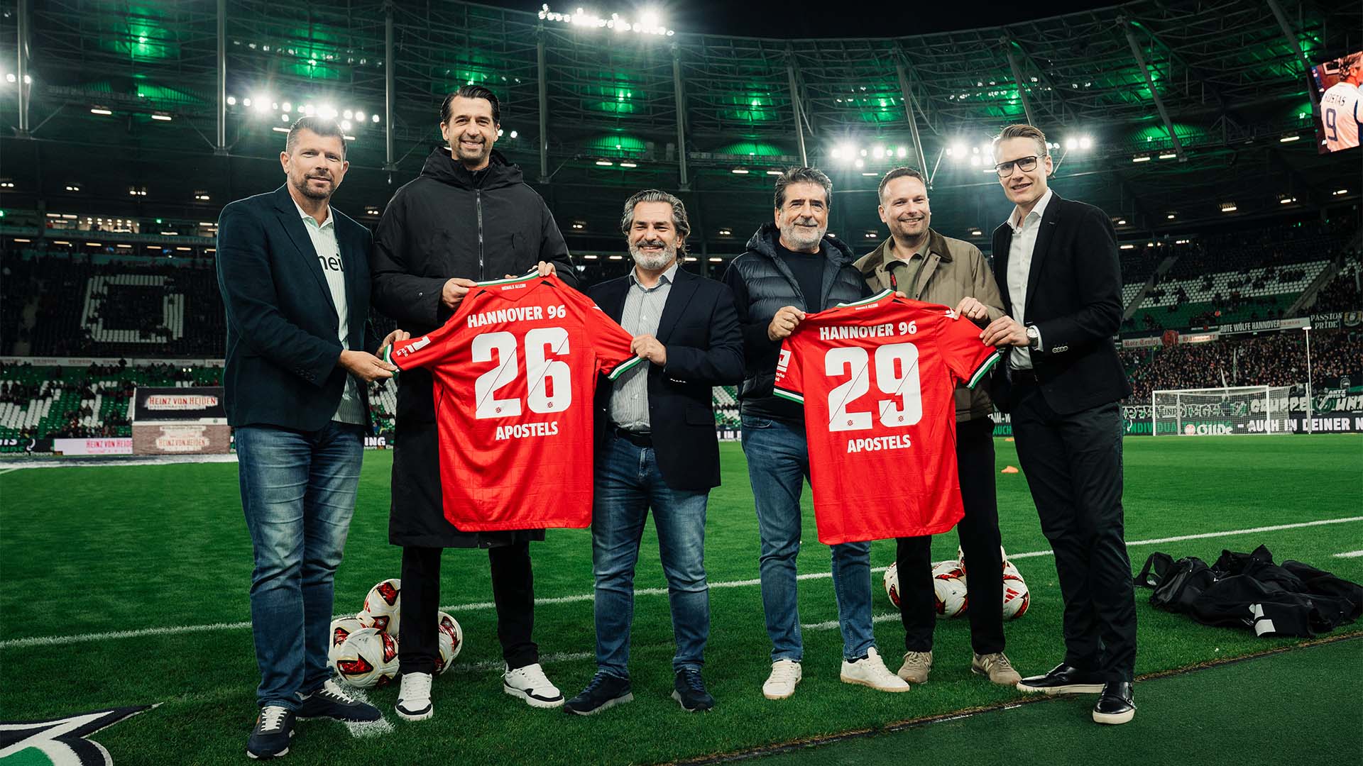 Sechs Männer posieren bei Flutlicht im 96-Stadion, jeweils drei von ihnen halten ein 96-Heimtrikot - eines mit der 26, eines mit der 29. 