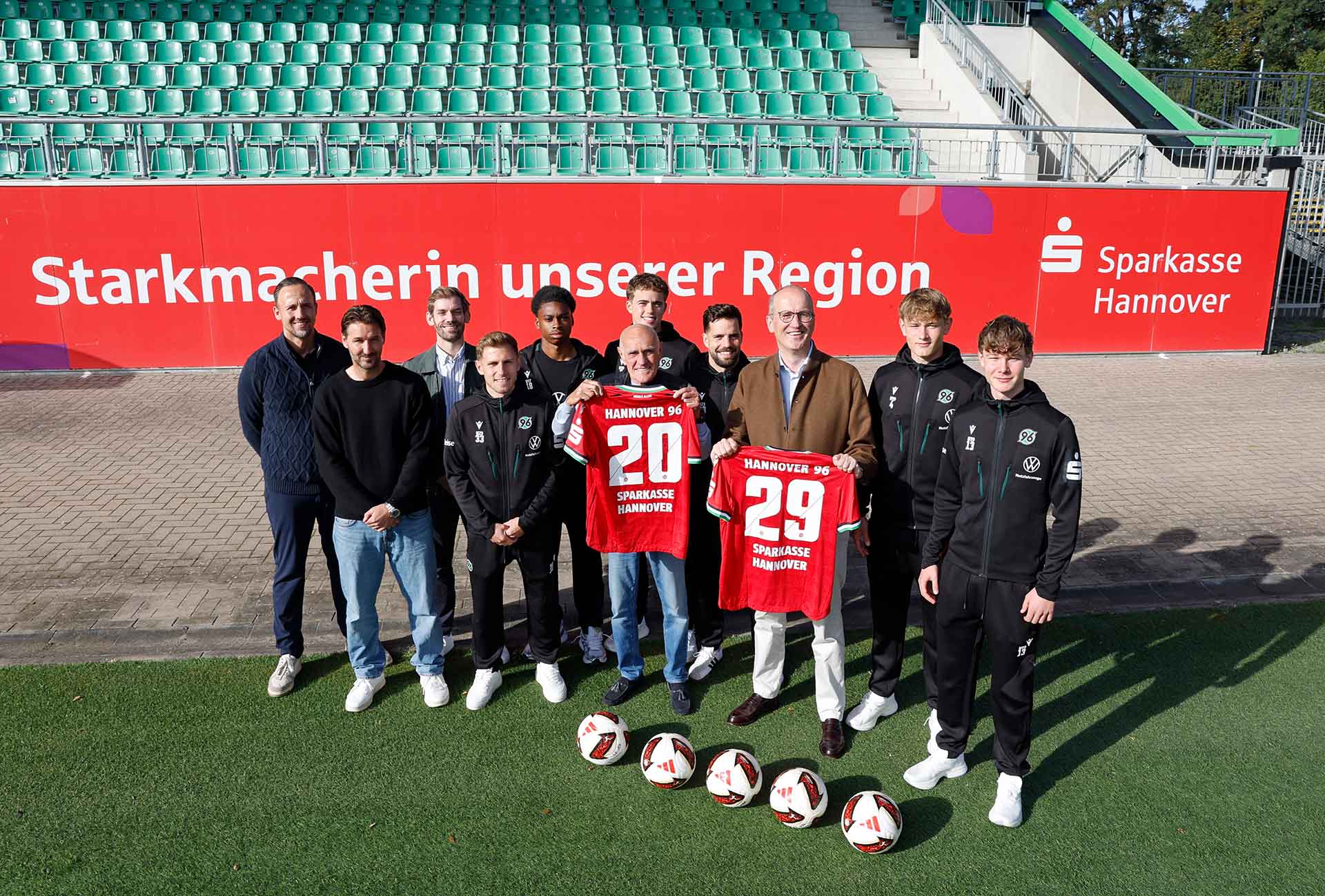 Eine Gruppe von elf jungen und älteren Männern steht auf dem Rasen des Eilenriedestadions, zwei halten 96-Heimtrikots mit dem Flock 29 und Sparkasse Hannover hoch. 