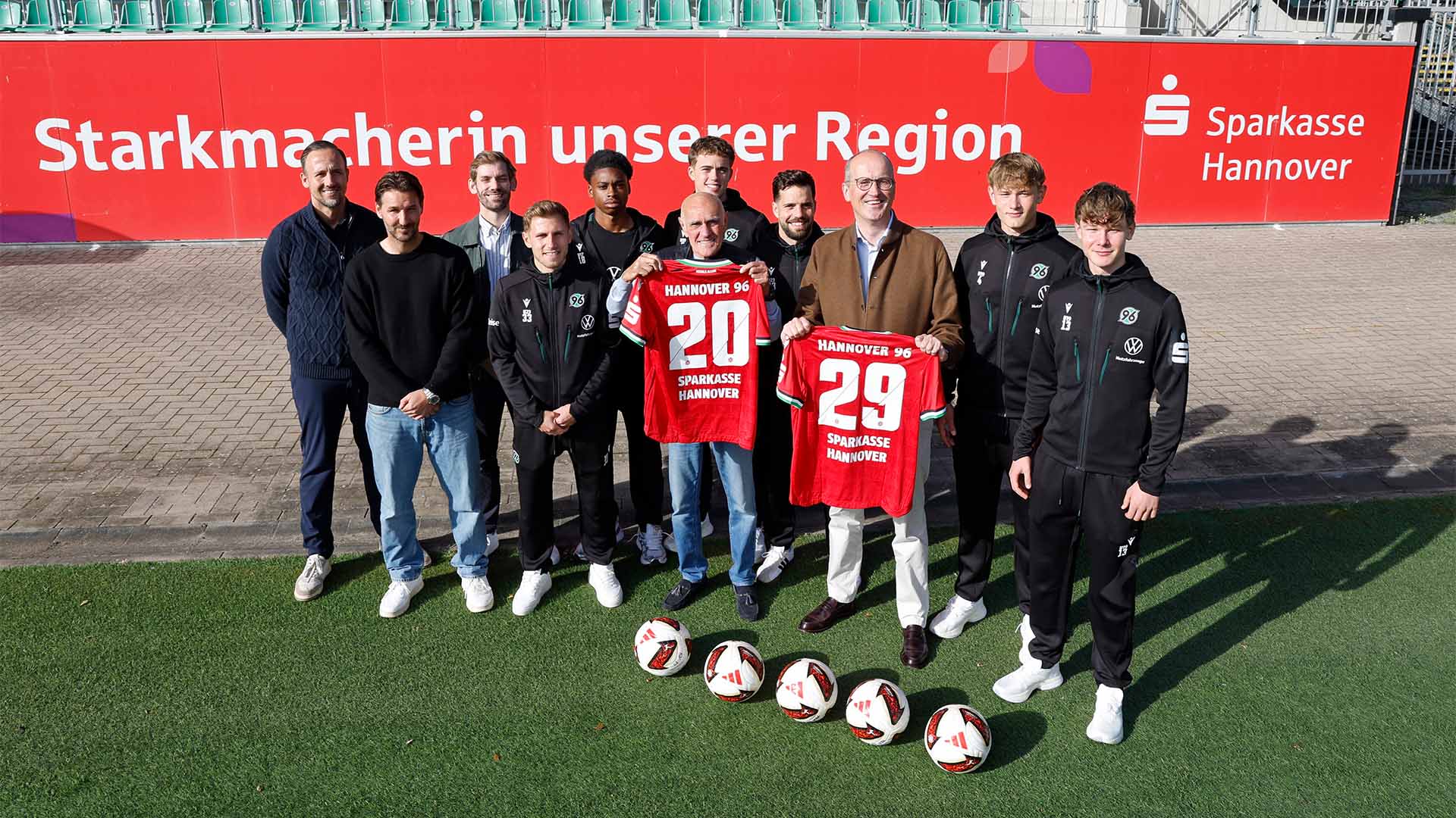 Eine Gruppe von elf jungen und älteren Männern steht auf dem Rasen des Eilenriedestadions, zwei halten 96-Heimtrikots mit dem Flock 29 und Sparkasse Hannover hoch. 