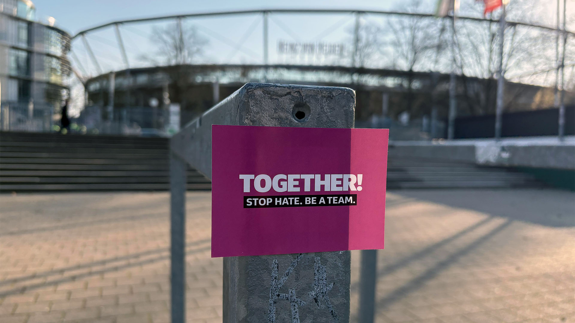 Ein pinker Sticker mit der Aufschrift "Together. Stop Hate. Be a Team." klebt an einem Pfosten, im Hintergrund unscharf das Stadion.