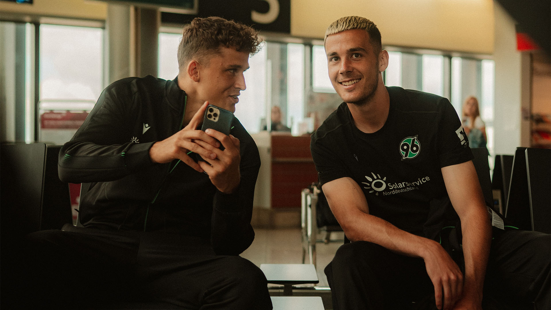 Benedikt Pichler und Bastian Allgeier warten am Flughafen, Pichler zeigt etwas am Handy, Allgeier lächelt in die Kamera.