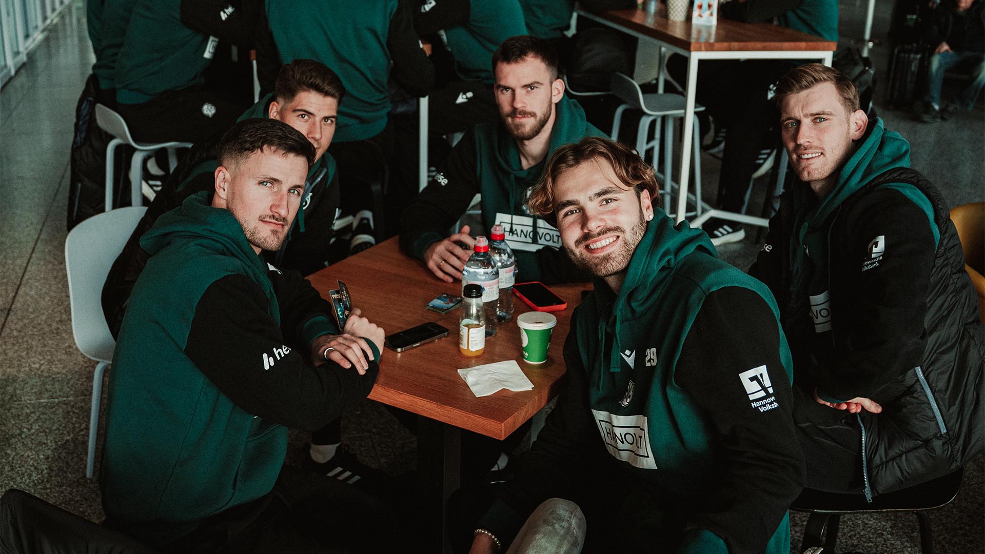 Enzo Leopold, Kolja Oudenne, Leo Weinkauf, Boris Tomiak und Franz Roggow sitzen am Flughafen an einem Tisch.