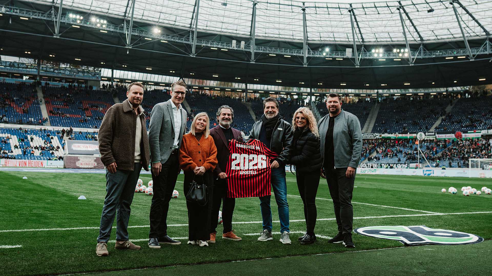 Verantwortliche von Hannover 96 und APOSTELS halten ein 96-Trikot im Stadion hoch.