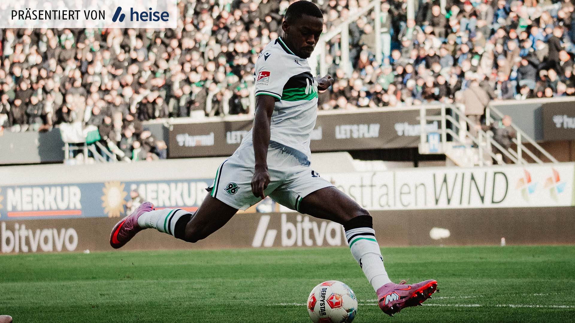 Noel Aseko im weißen 96-Ausweichtrikot holt zum Torschuss aus, während der Ball vor ihm auf dem Rasen liegt.