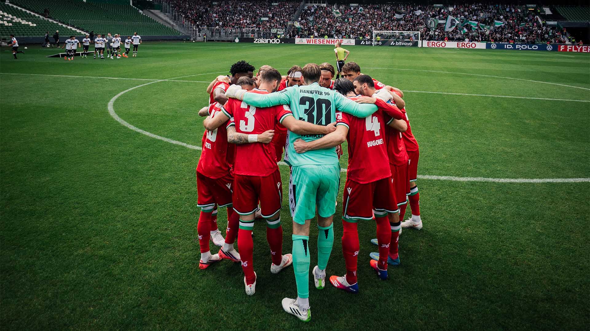 Die 96-Mannschaft steht in einem Kreis auf dem Rasen im Weserstadion, im Hintergrund die 96-Fankurve.