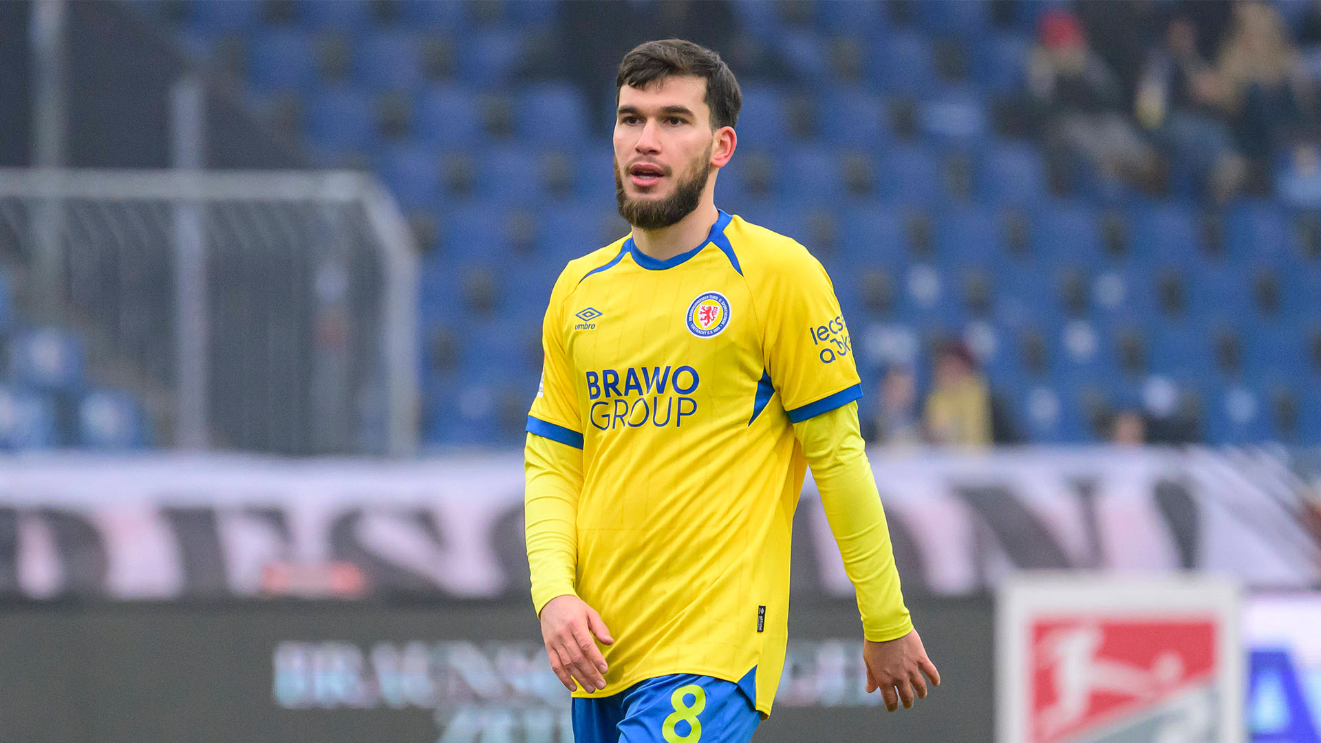 Mehmet Aydin in einem gelben Trikot geht über den Rasen. Auf seiner blauen Hose ist die Nummer 8 in gelber Farbe zu sehen. Die Zuschauertribüne im Hintergrund ist unscharf.