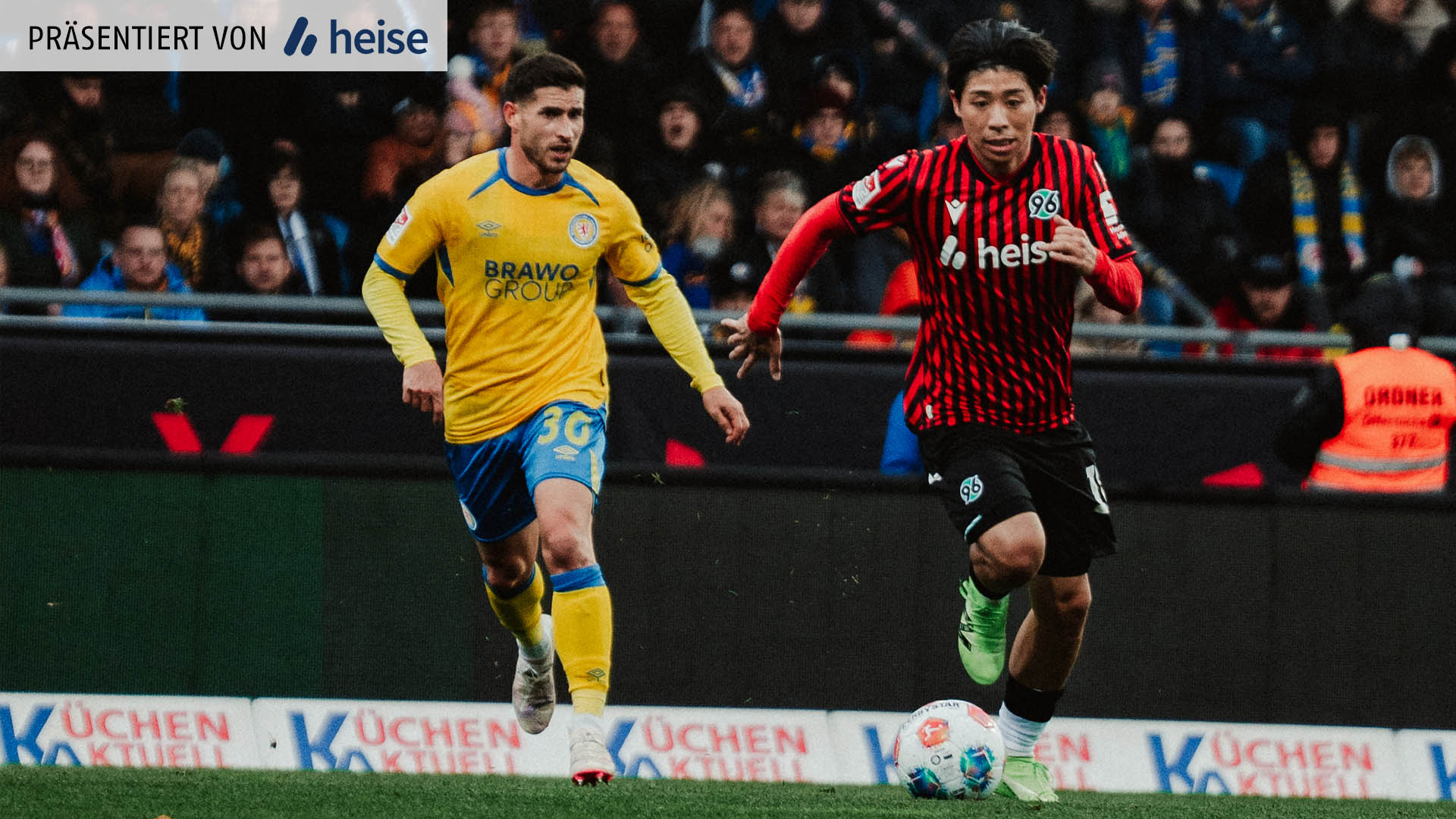 Daisuke Yokota im rot-schwarzen Trikot läuft mit dem Ball über den Rasen, während ein Gegenspieler im gelb-blauen Trikot dicht hinter ihm herläuft. Im Hintergrund sind Zuschauer auf der Tribüne zu sehen.