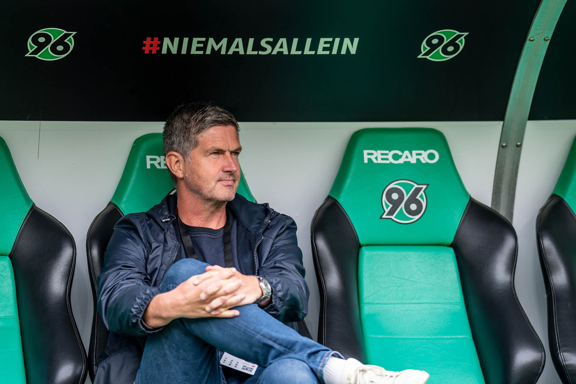 Ralf Becker sitzt auf der Ersatzbank im Stadion, hat das rechte Bein auf dem linken Knie und die Hände davor verschränkt, schaut leicht nach links.  