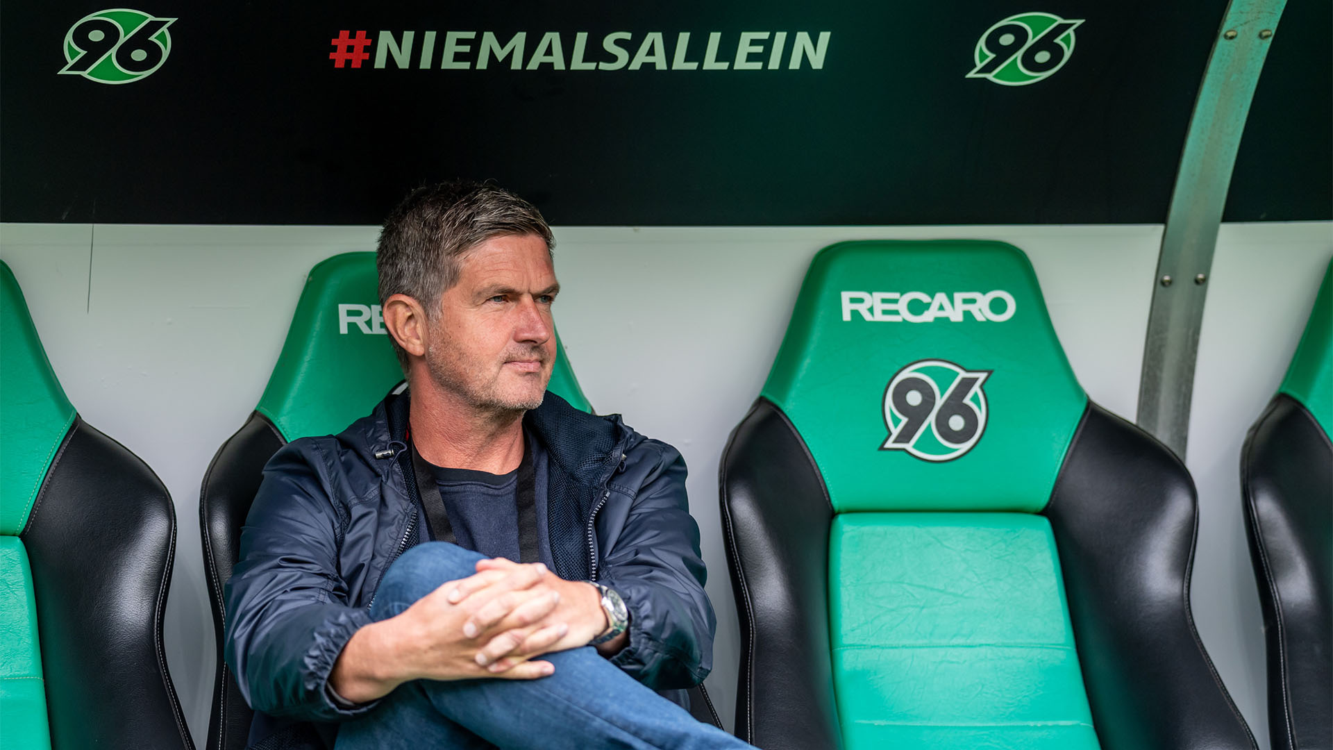 Ralf Becker sitzt auf der Ersatzbank im Stadion, hat das rechte Bein auf dem linken Knie und die Hände davor verschränkt, schaut leicht nach links.  