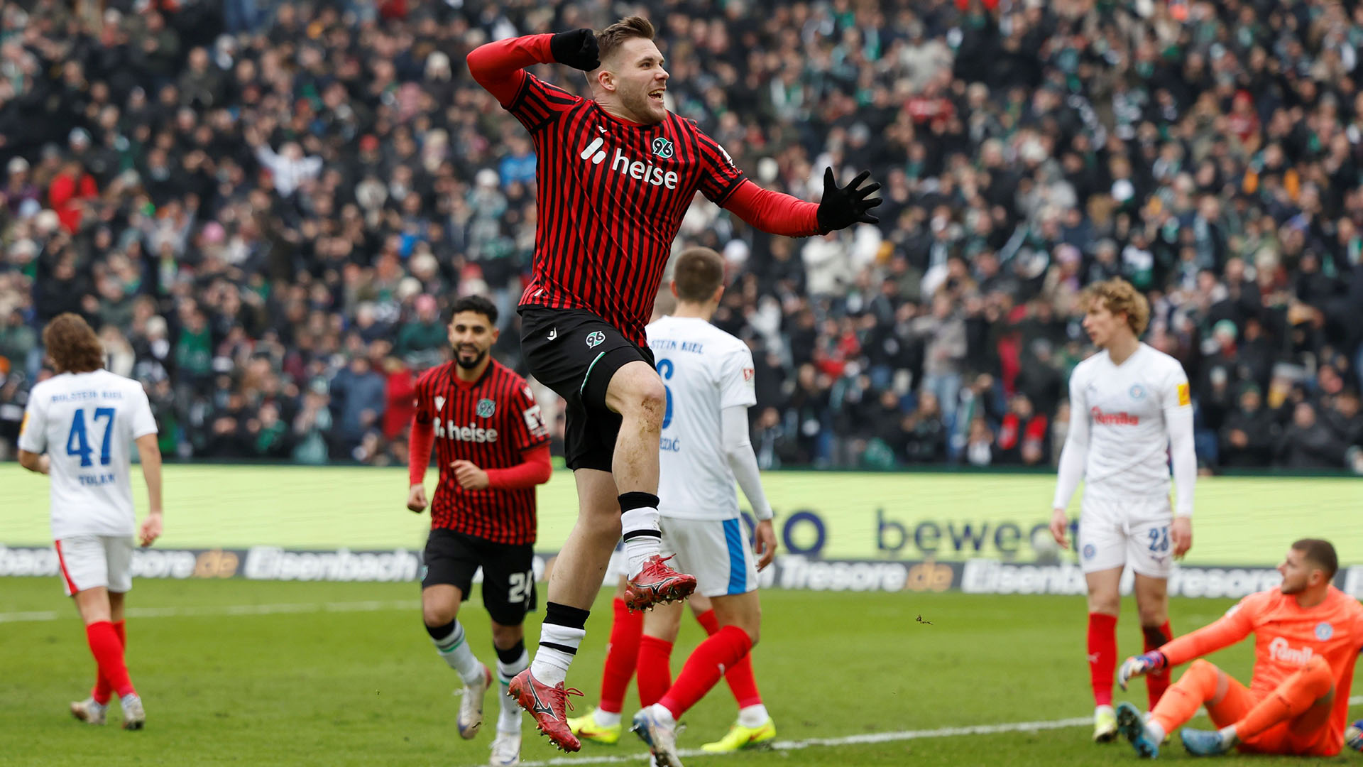 Benjamin Källman springt jubelnd in die Luft. Er trägt ein rot-schwarz gestreiftes Trikot und im Hintergrund sind Spieler von Holstein Kiel sowie der Torwart am Boden zu sehen.