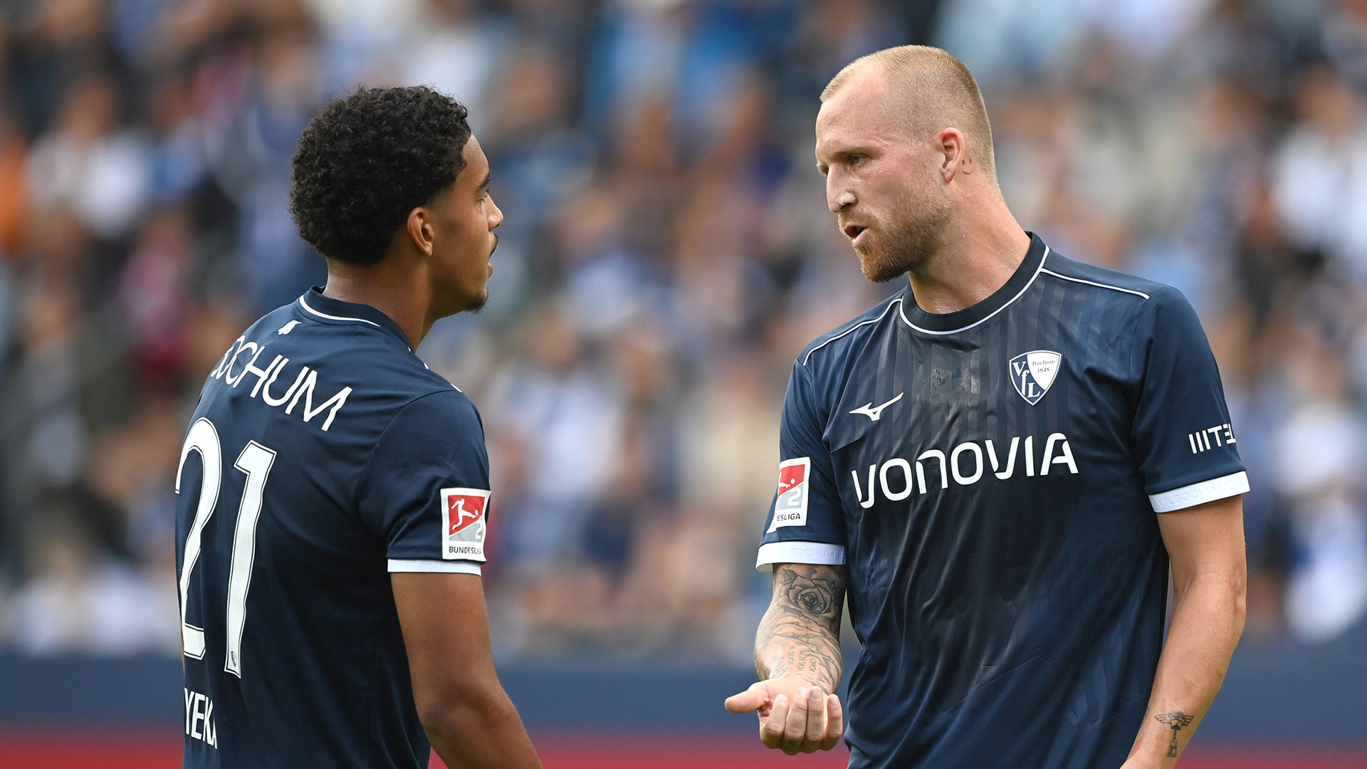 Francis Onyeka und Philipp Hofmann vom VfL Bochum tragen dunkelblaue Trikots und sprechen auf dem Platz miteinander. Im Hintergrund sind unscharf die Zuschauerränge zu sehen.