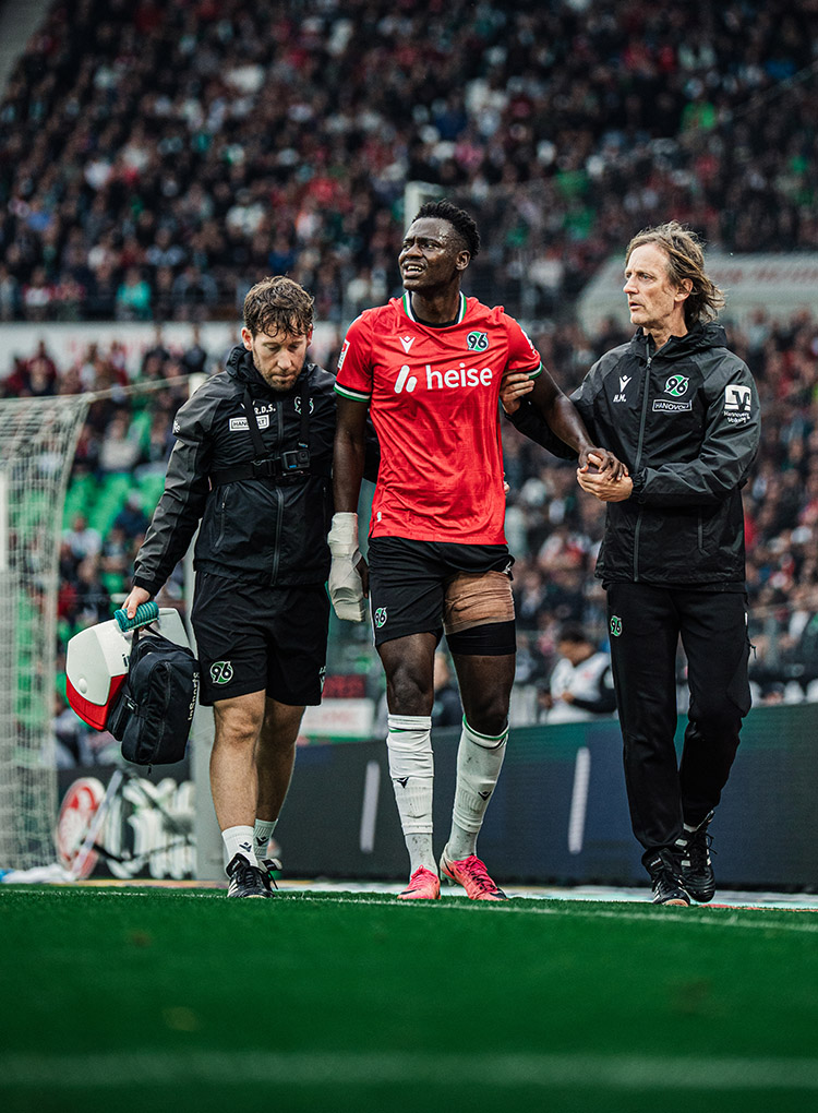 Der 96-Spieler Mustapha Bundu wird von einem 96-Physiotherapeuten und dem 96-Mannschaftsarzt vom Platz geführt, sein linker Oberschenkel ist bandagiert.