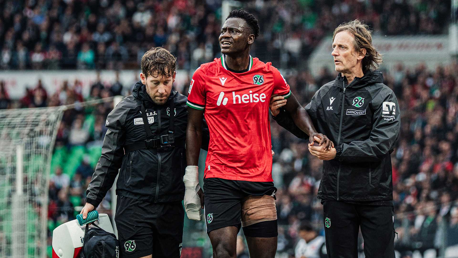 Der 96-Spieler Mustapha Bundu wird von einem 96-Physiotherapeuten und dem 96-Mannschaftsarzt vom Platz geführt, sein linker Oberschenkel ist bandagiert.