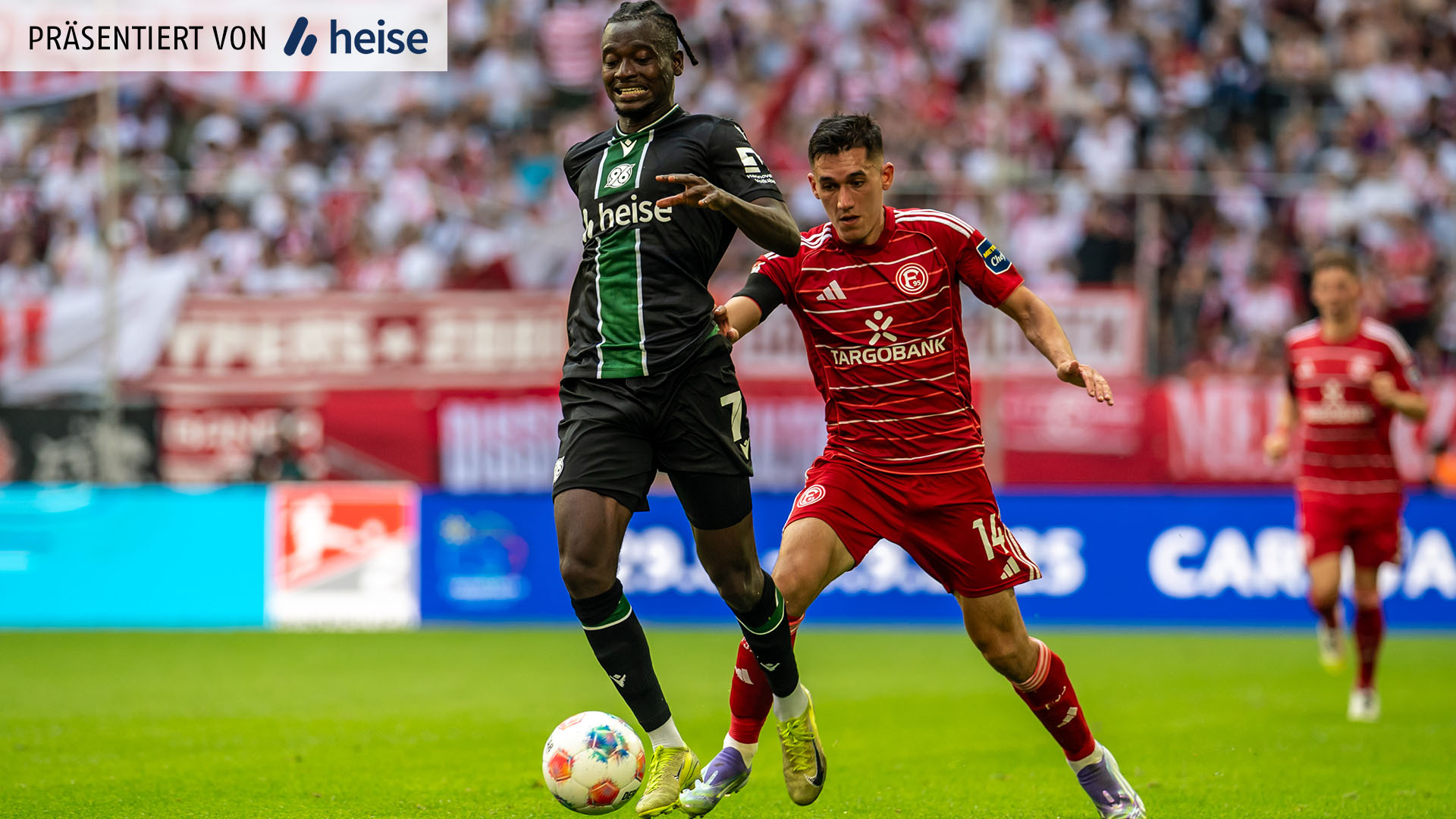 Ein Spieler von Hannover 96 im schwarzen Auswärtstrikot kontrolliert den Ball, während ein Düsseldorfer im roten Trikot dicht hinter ihm läuft und den Arm ausstreckt.