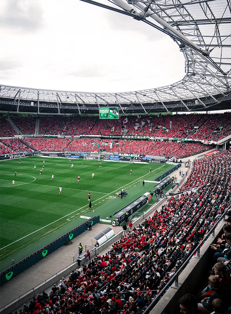 Blick in Richtung Nordkurve im vollen Stadion, die meisten Fans tragen rote Sachen, es läuft gerade ein Spiel.