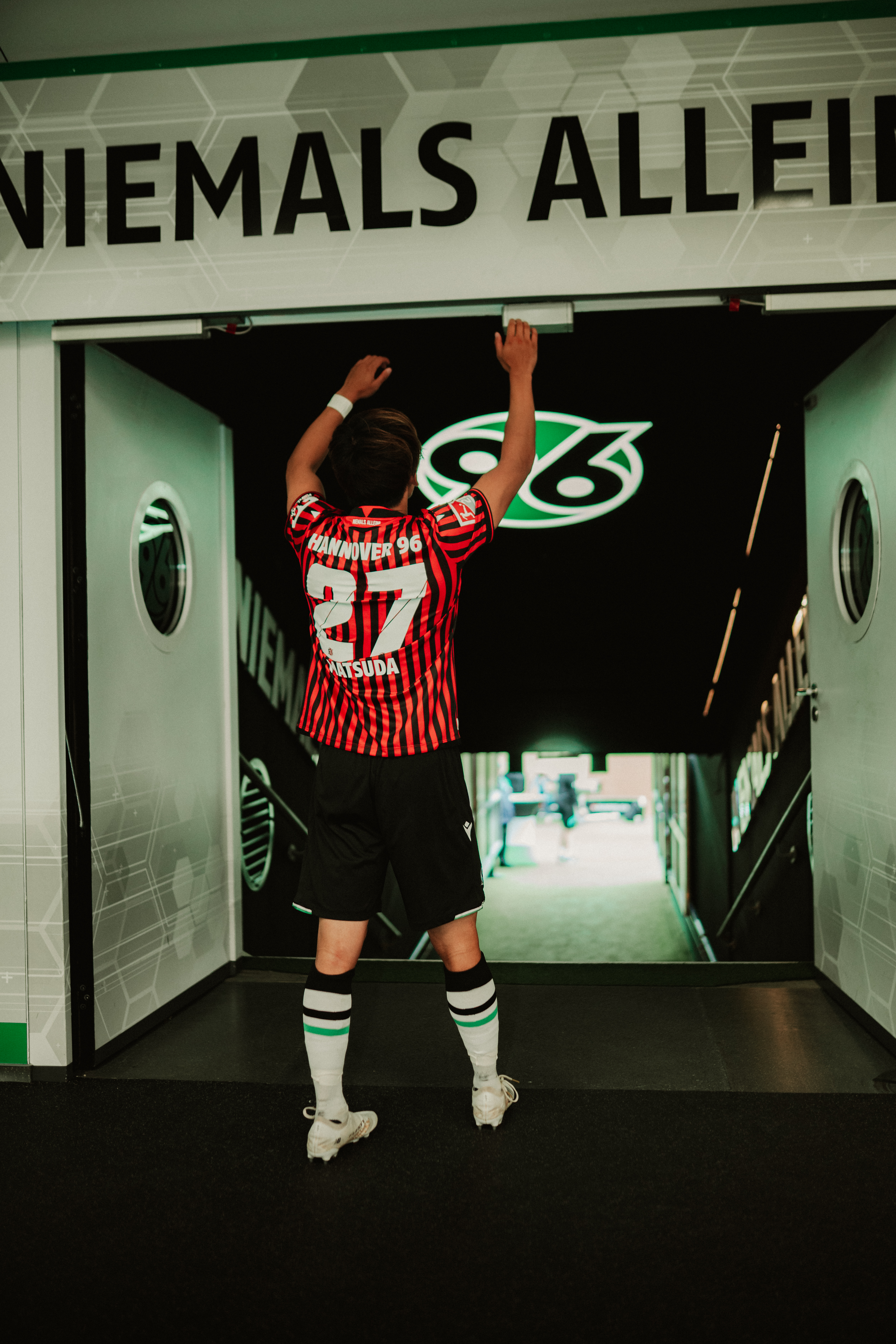 Matsuda vor dem Spielertunnel mit den Händen. vor einem "Niemals Allein" Schild.