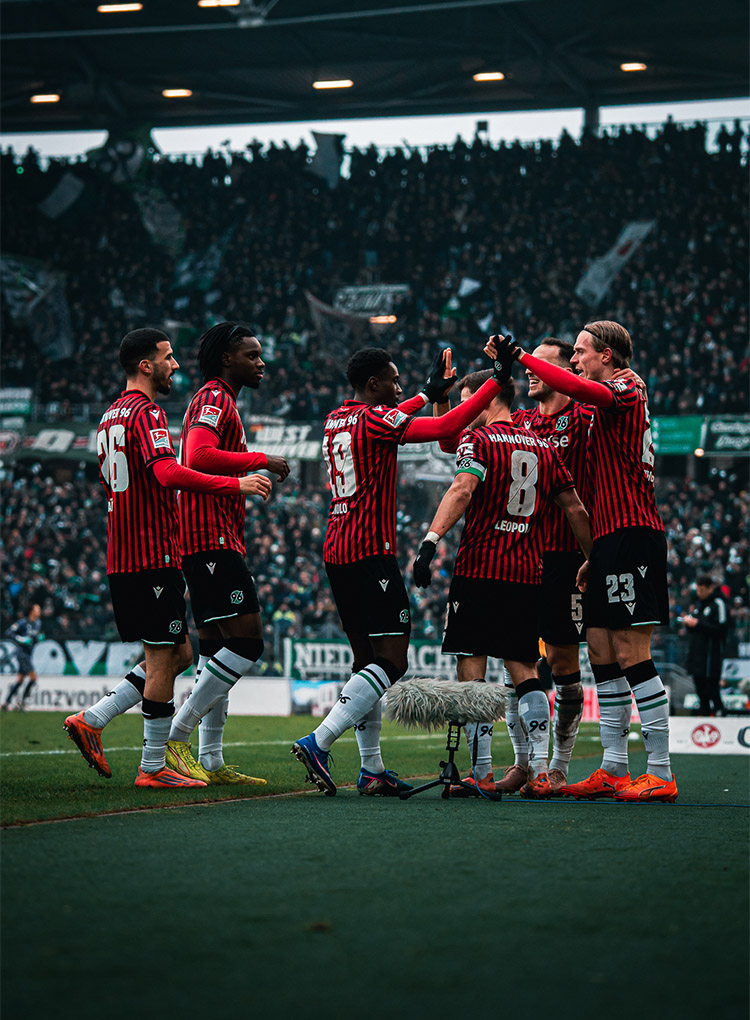 Sechs 96-Spieler in Heimtrikots bejubeln ein Tor, im Hintergrund die volle Nordkurve.