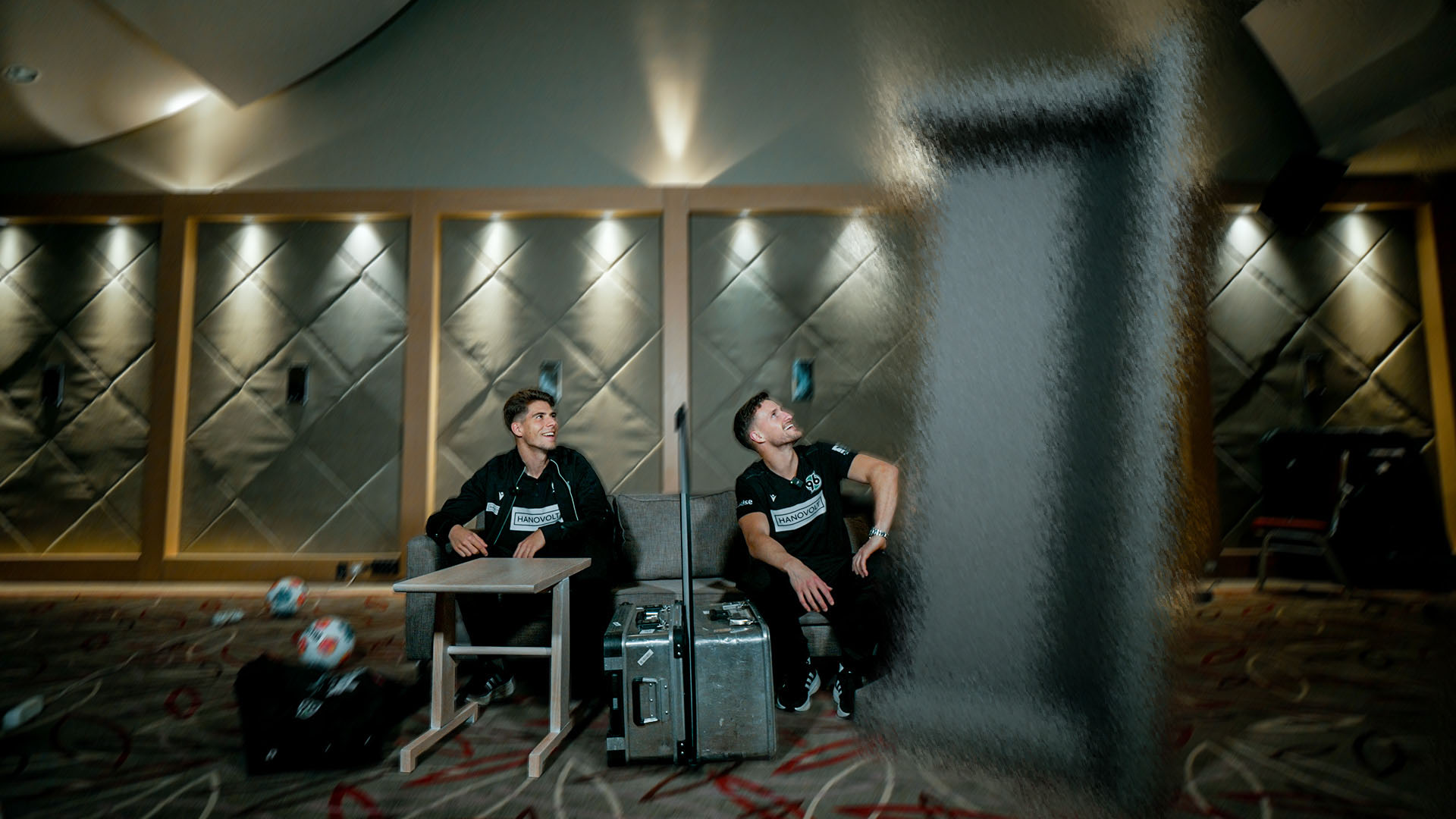 Franz Roggow und Enzo Leopold sitzen mit einer kleinen Trennwand zwischen sich und schauen auf einen großen Gegenstand neben Enzo, der geblurrt ist.