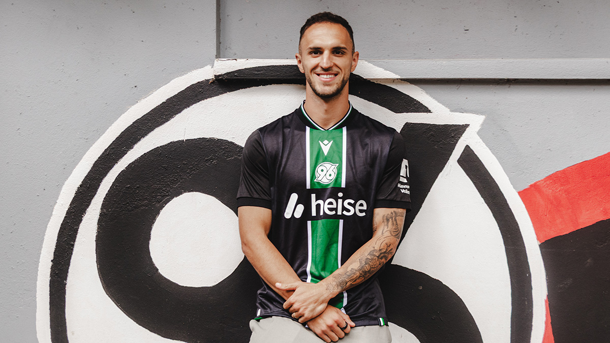 Virgil Ghita lehnt mit dem Rücken an einer Wand, auf dem ein Logo von 96 prangt. 