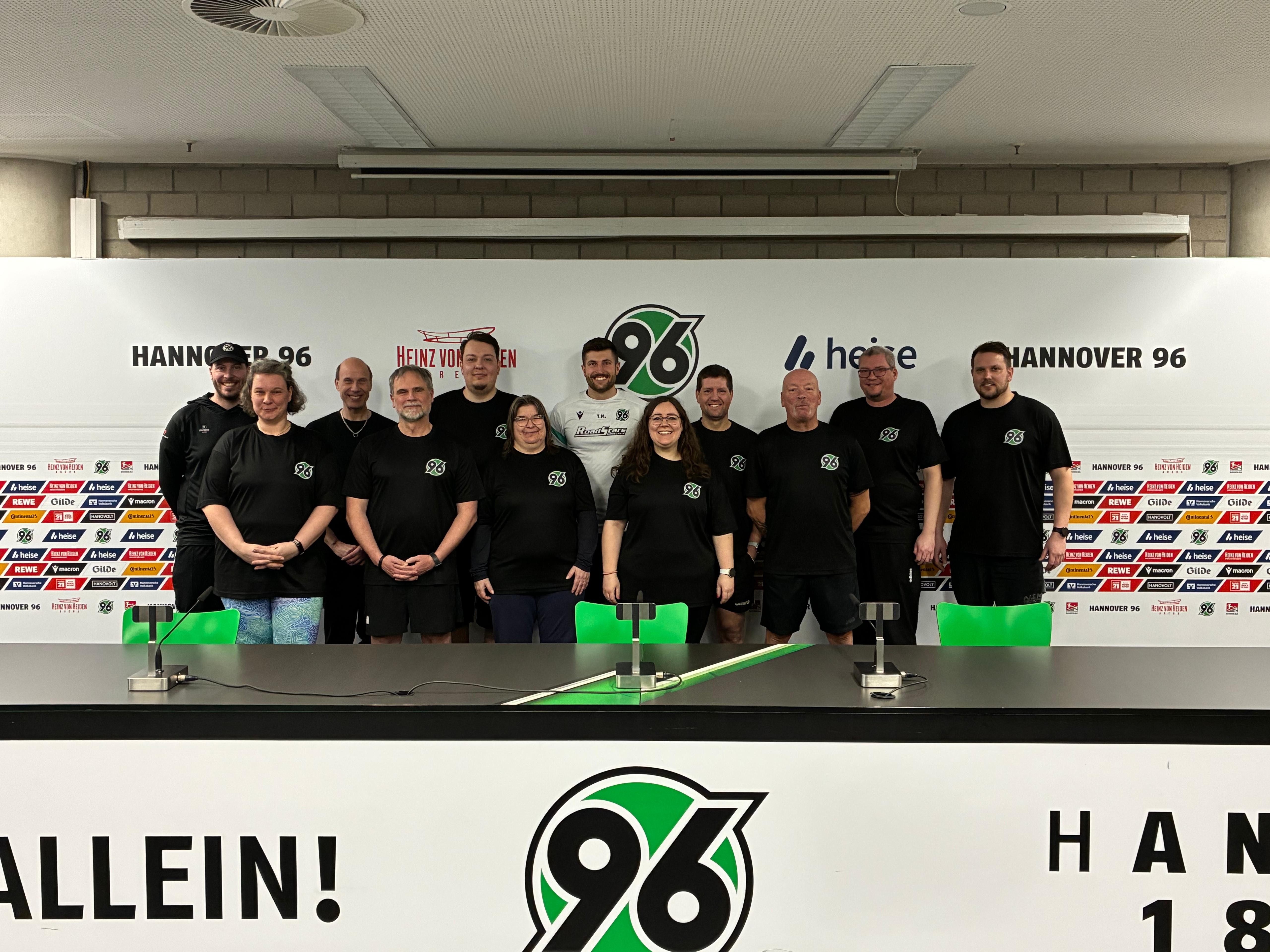 Eine Gruppe von Menschen in schwarze 96-T-Shirts steht auf dem Podium des Pressekonferenzraums. 