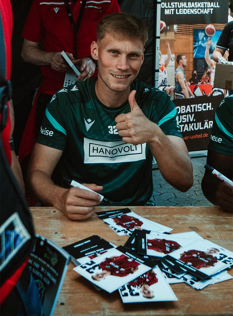 Jonas Sterner zeigt den linken Daumen hoch und lächelt, vor ihm liegt ein Stapel Autogrammkarten.