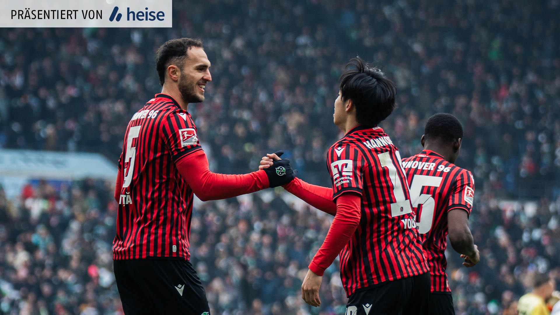Zwei Spieler von Hannover 96 in rot-schwarz gestreiften Trikots geben sich nach einem Treffer die Hand und schauen sich dabei in die Augen. Im unscharfen Hintergrund sind die Zuschauerränge zu sehen.
