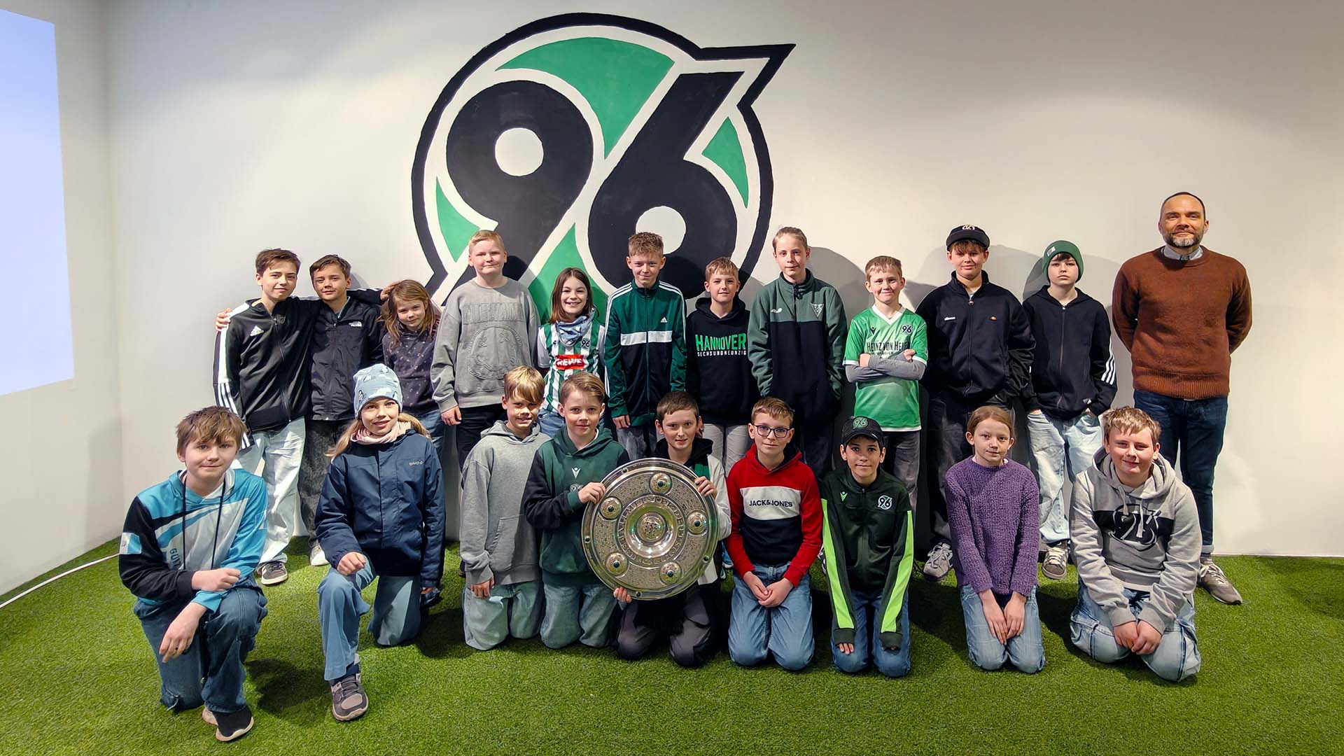 Die Kindergruppe posiert mit der Meisterschale vor einem großen 96-Logo.