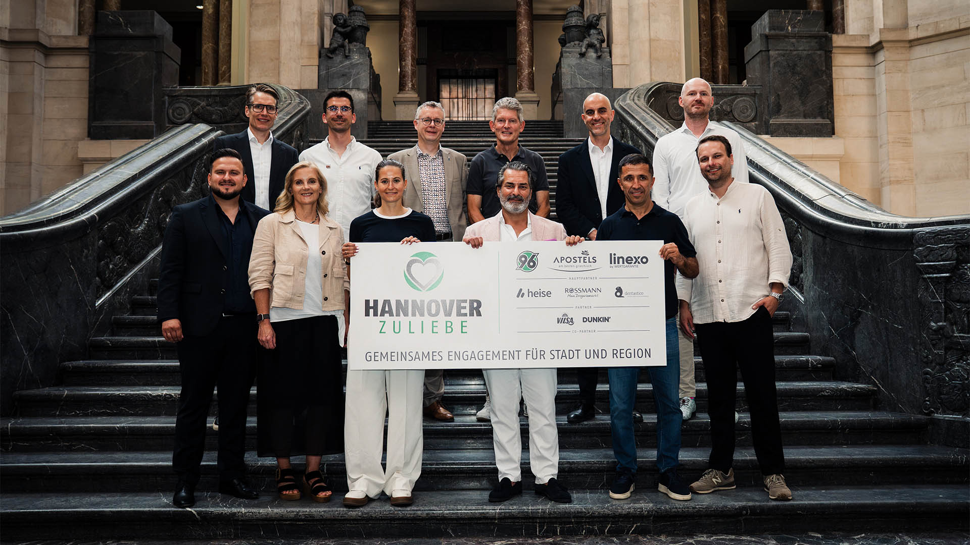Zwölf Personen stehen in zwei Reihen auf einer Treppe. Sie halten ein Schild mit der Aufschrift "HANNOVER ZULIEBE" und einigen Logos drauf.