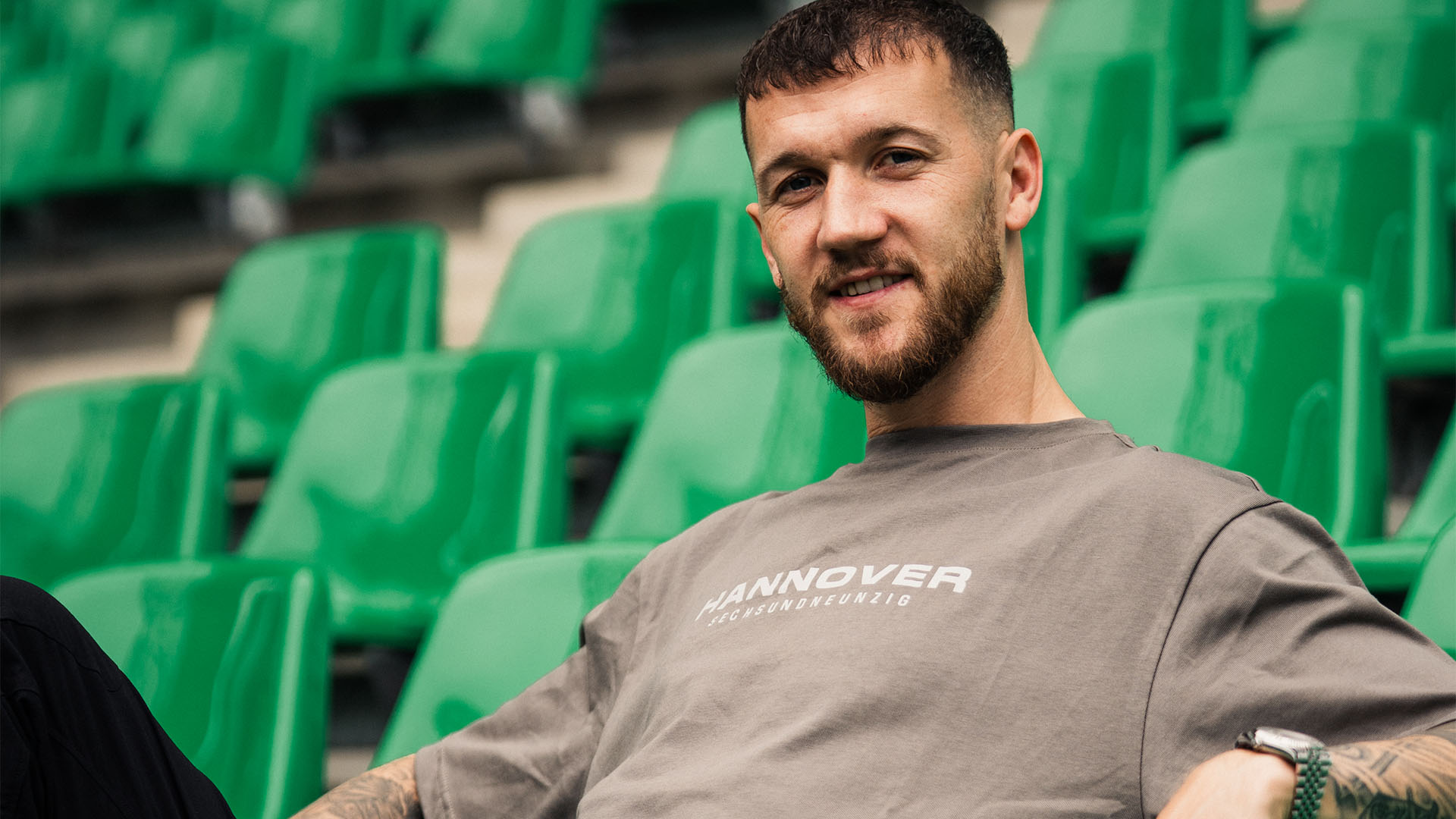 Marcel Hartel sitzt in einem hellbraunen 96-T-Shirt auf grünen Stadionsitzen.
