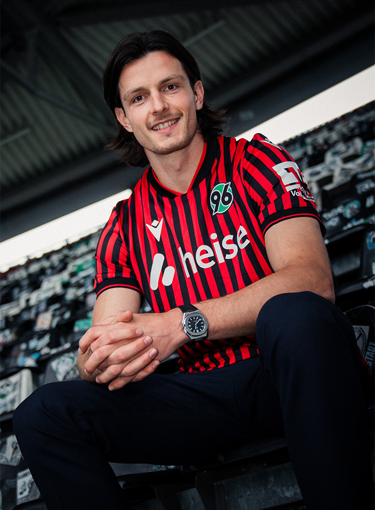 Jean Hugonet sitzt m 96-Heimtrikot auf der Tribüne der Heinz von Heiden Arena und lächelt in die Kamera.
