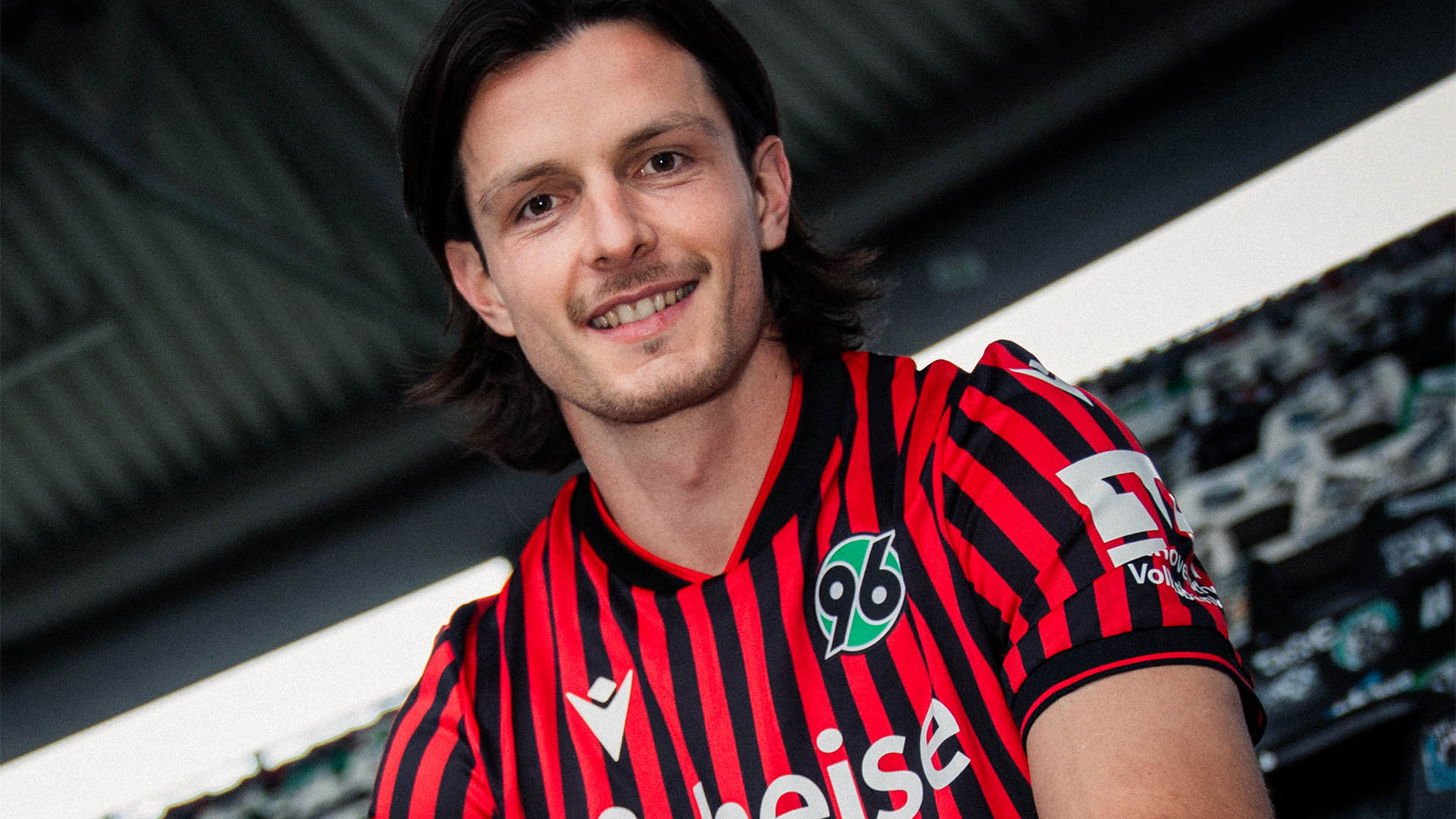 Jean Hugonet sitzt m 96-Heimtrikot auf der Tribüne der Heinz von Heiden Arena und lächelt in die Kamera.