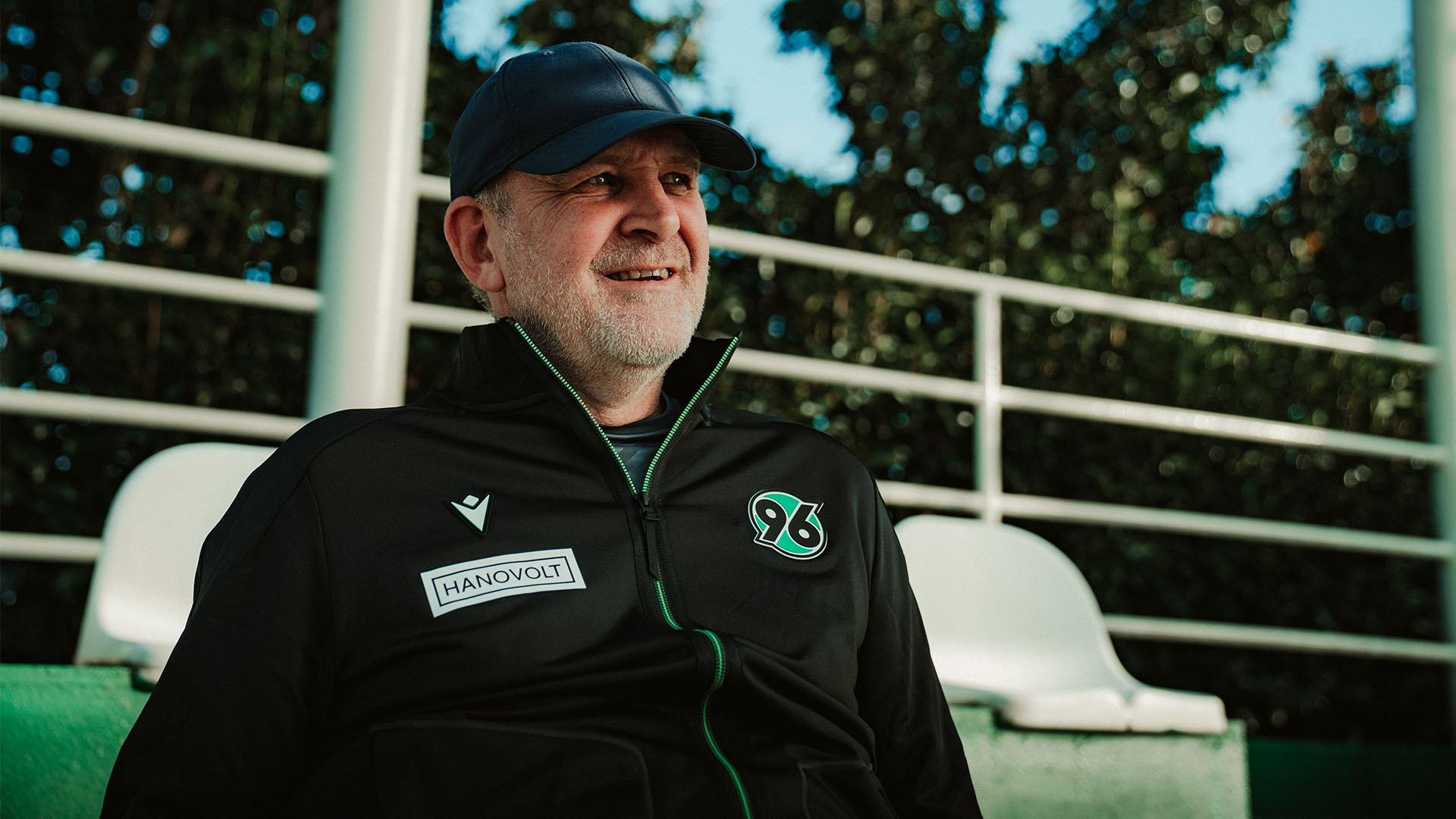 Jörg Schmadtke mit 96-Jacke und Cap schaut lächelnd zur Seite.