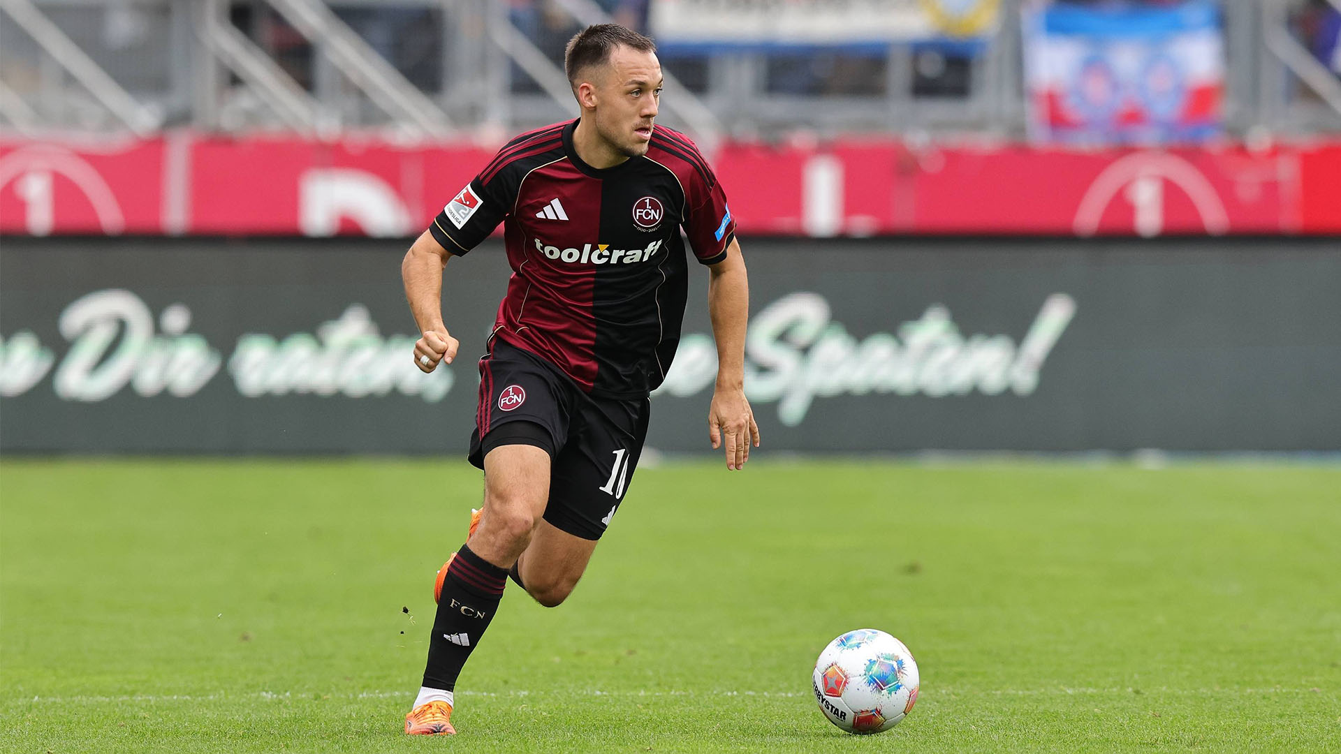 Julian Justvan im schwarz-roten Heimtrikot des 1. FC Nürnbergs läuft über den Platz mit dem Ball vor den Füßen und schaut über seine linke Schulter.