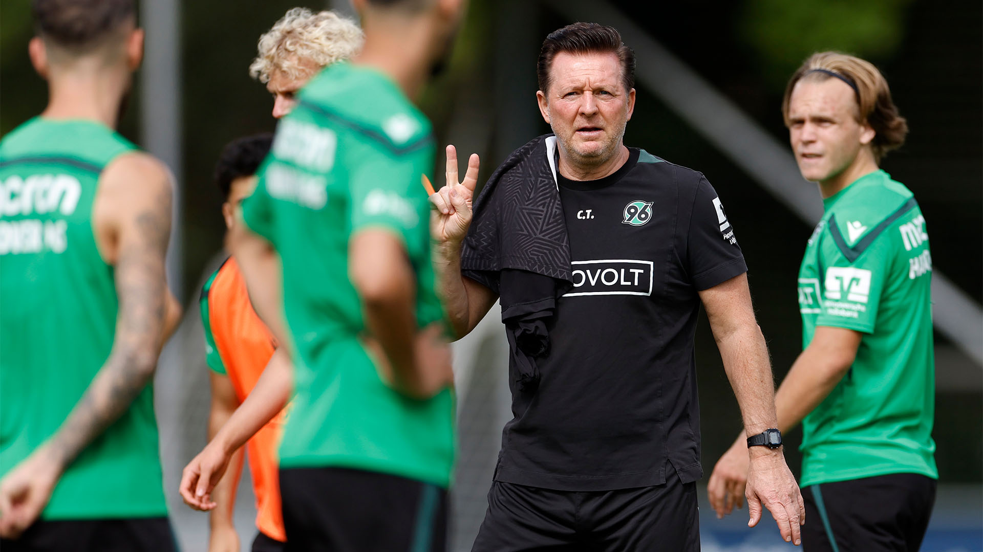 96-Cheftrainer Christian Titz zeigt mit zwei Fingern der rechten Hand, um ihn herum 96-Spieler. 