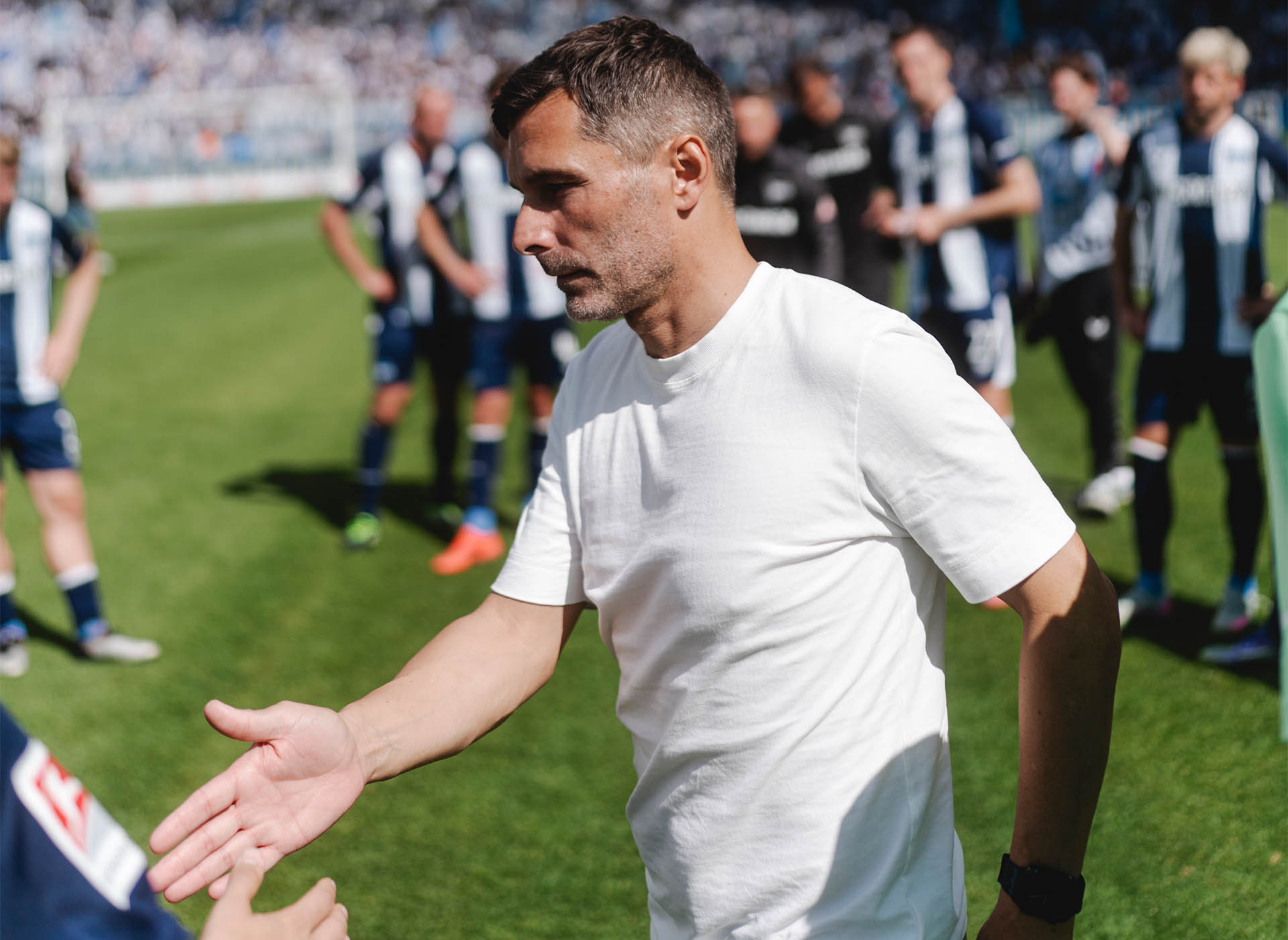 Stefan Leitl klatscht im Olympiastadion mit einem nicht erkennbaren Spieler ab, die Sonne scheint, er trägt ein weißes T-Shirt.