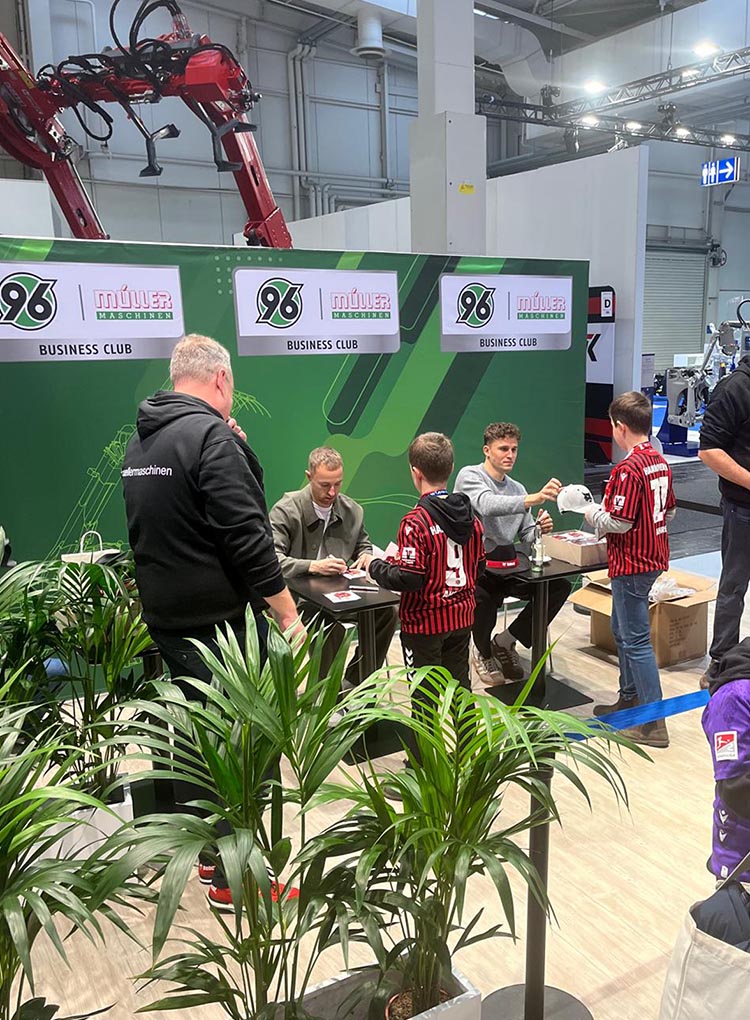 Maurice Neubauer und Benedikt Pichler sitzen an Tischen am Agritechnica-Stand von Müller Maschinen und schreiben Autogramm für zwei Kinder.
