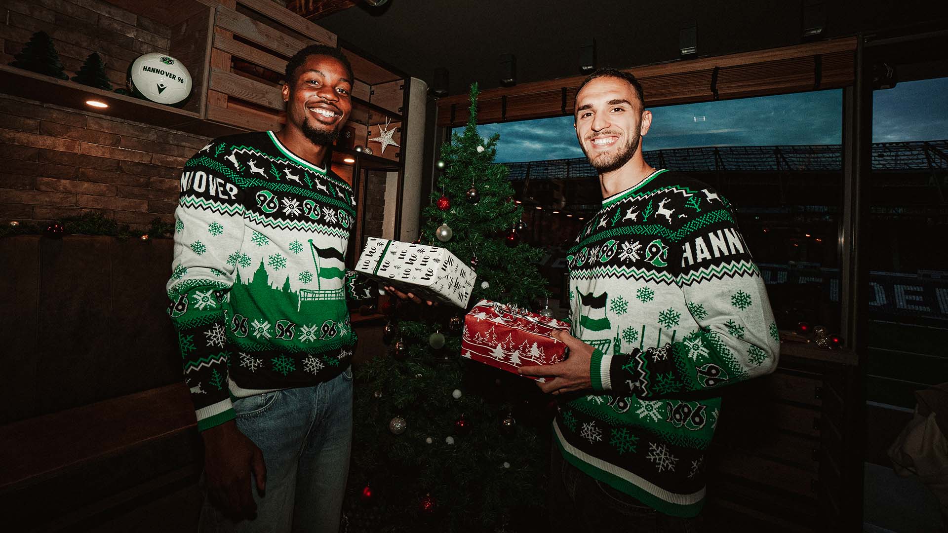 Ime Okon und Virgil Ghita tragen den 96-Weihnachtspulli, strahlen in die Kamera und halten Geschenke in den Händen. Im Hintergrund ein Weihnachtsbaum.