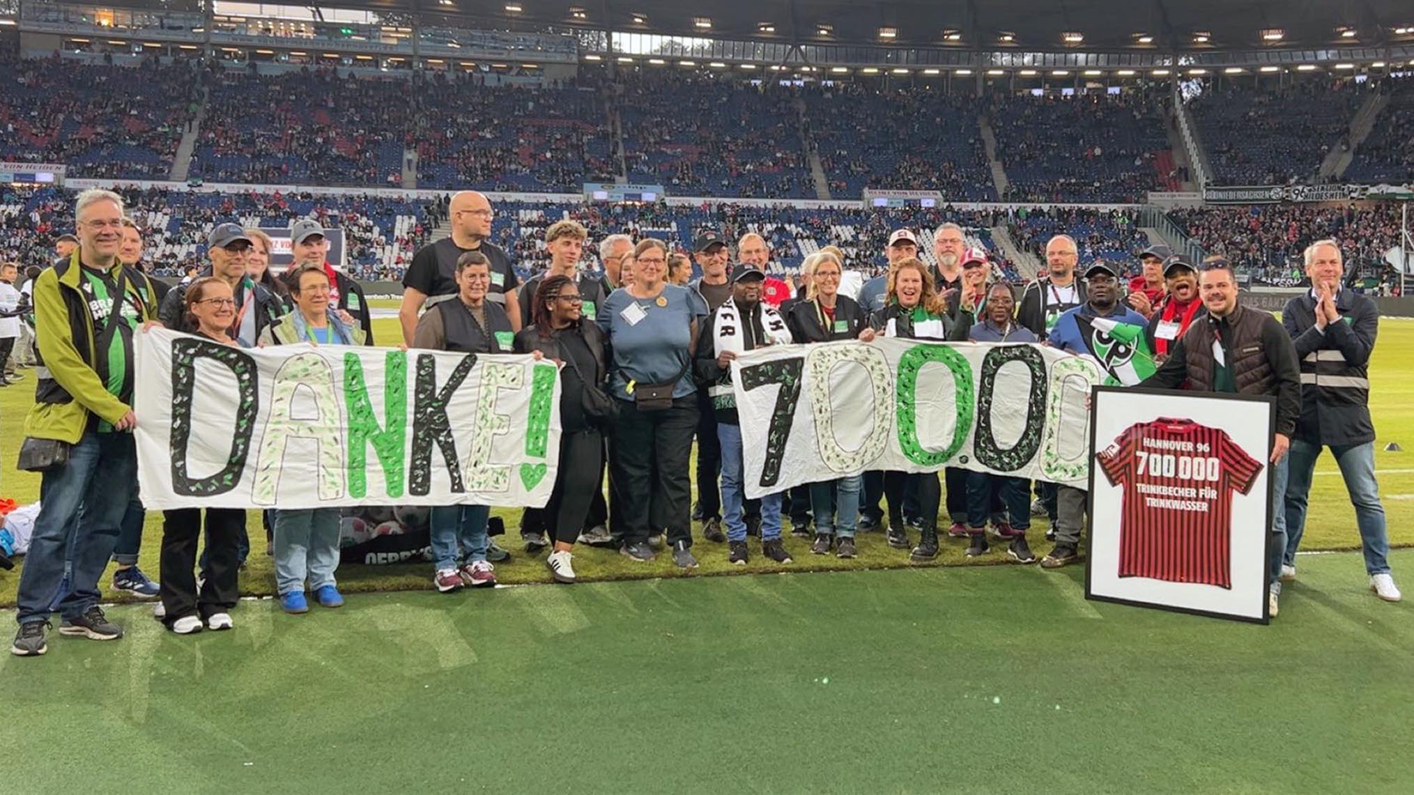 Das Trinkbecher-Team posiert im Stadion mit einem "Danke! 700.000"-Banner sowie einem eingerahmten Trikot. 