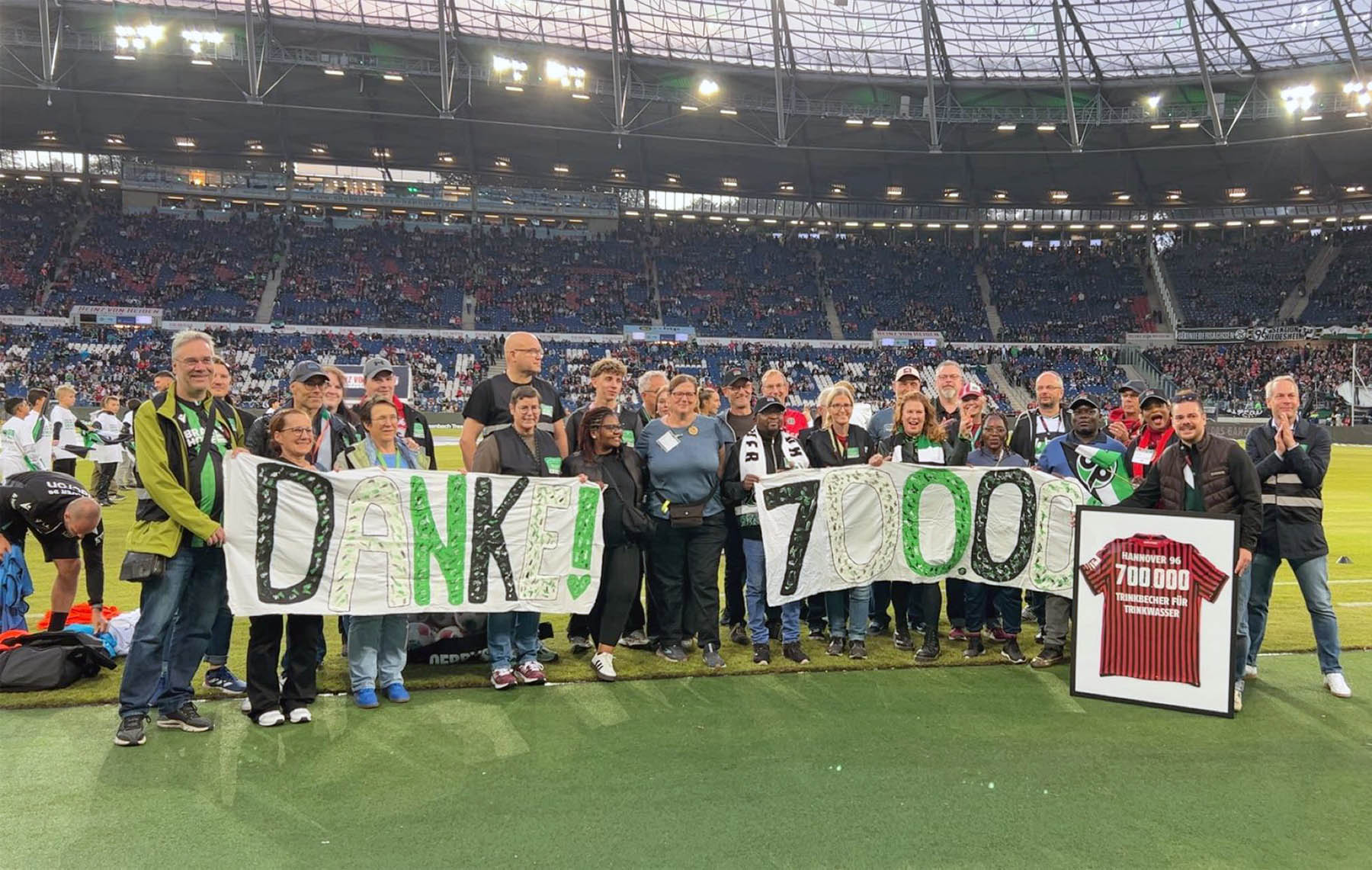 Das Trinkbecher-Team posiert im Stadion mit einem "Danke! 700.000"-Banner sowie einem eingerahmten Trikot. 