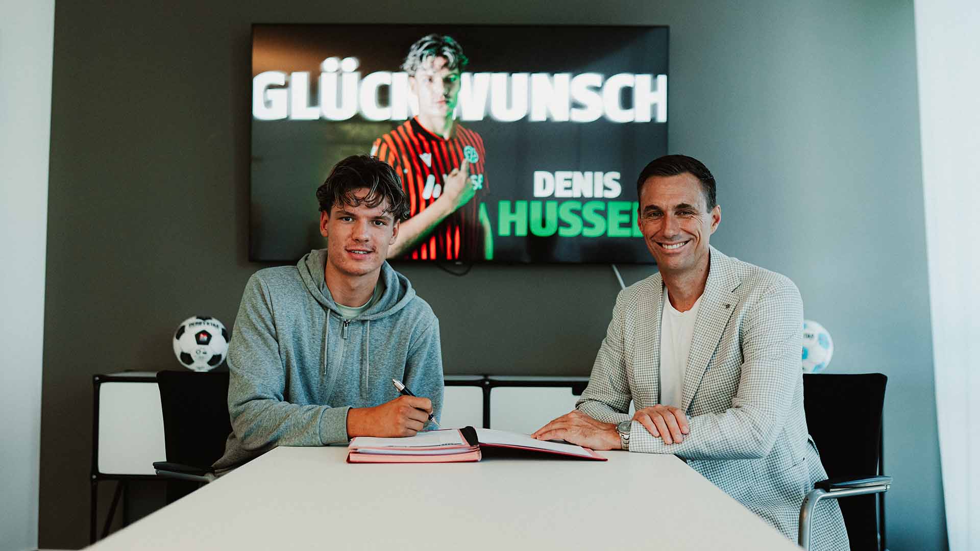 Denis Husser und Marcus Mann sitzen am Schreibtisch in Manns Büro, Husser unterschreibt seinen Vertrag, beide lächeln.