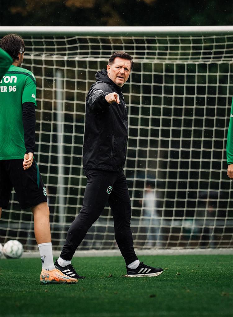 96-Cheftrainer Christian Titz steht auf dem Trainingsplatz und zeigt mit dem rechten Zeigefinger.