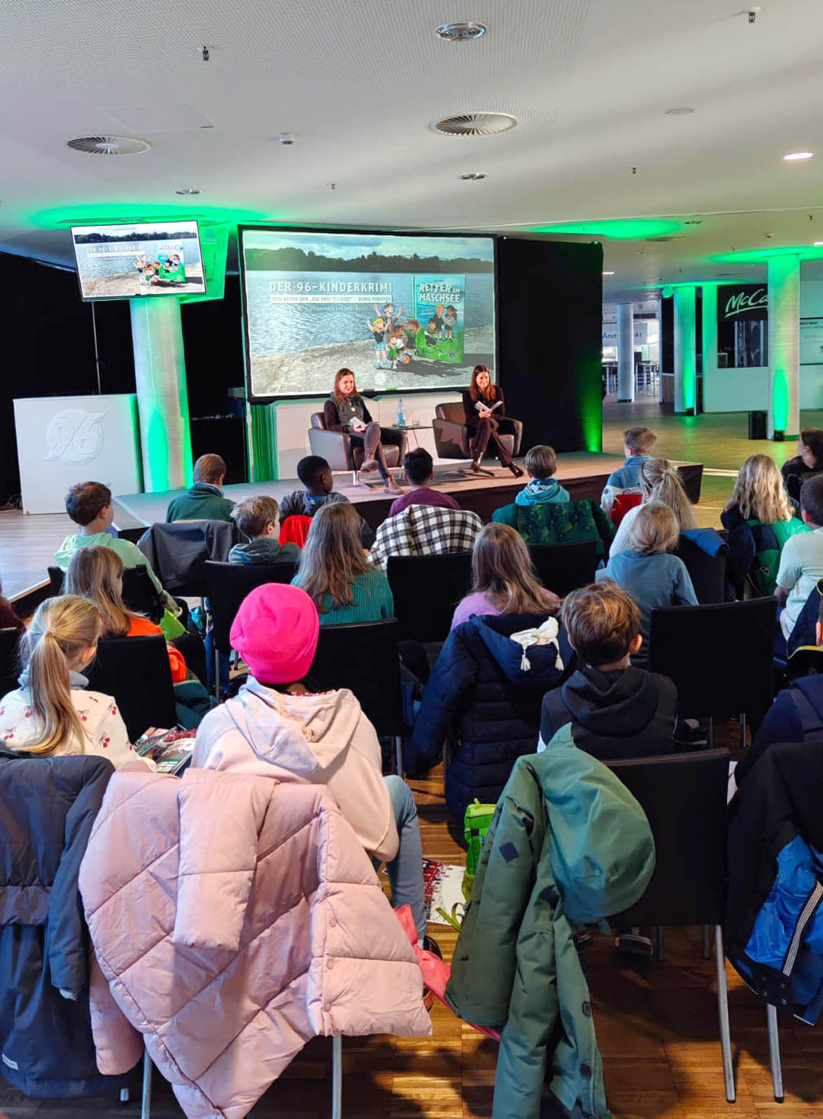 Johanna Heise und Sally Preiss aus dem KIDS-Team sitzen auf der Bühne und lesen vor. Davor sitzen die Kinder und hören zu.