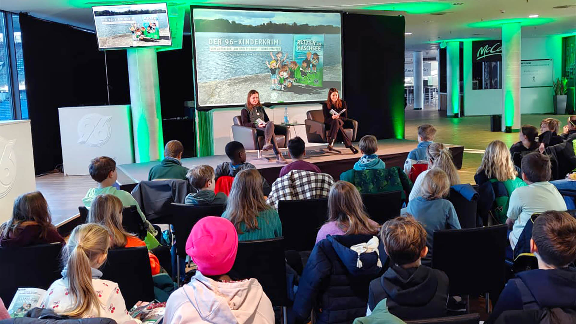 Johanna Heise und Sally Preiss aus dem KIDS-Team sitzen auf der Bühne und lesen vor. Davor sitzen die Kinder und hören zu.