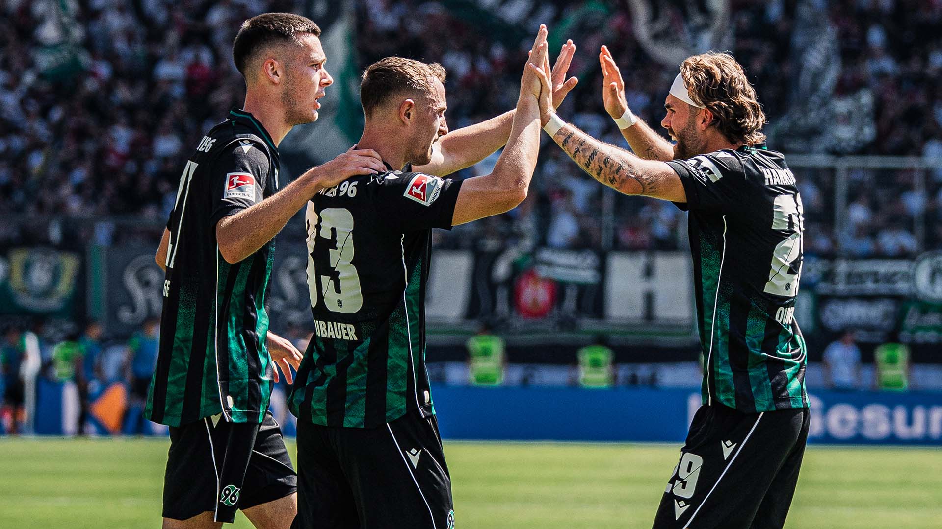 Drei 96-Spieler in den schwarz-grünen Auswärtstrikot klatschen miteinander ab, im Hintergrund ein voller Block mit 96-Fans.