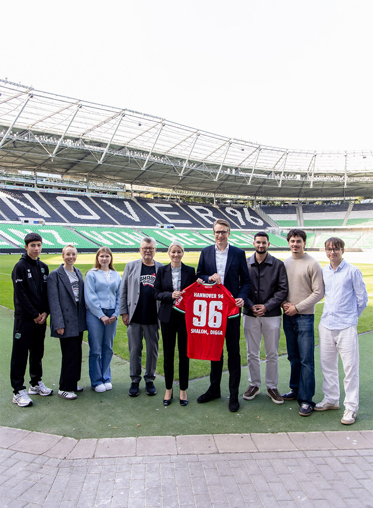 Eine Gruppe von neun Personen steht im 96-Stadion, zwei Personen halten ein 96-Heimtrikot mit der Aufschrift "Shalom, Digga" hoch. 