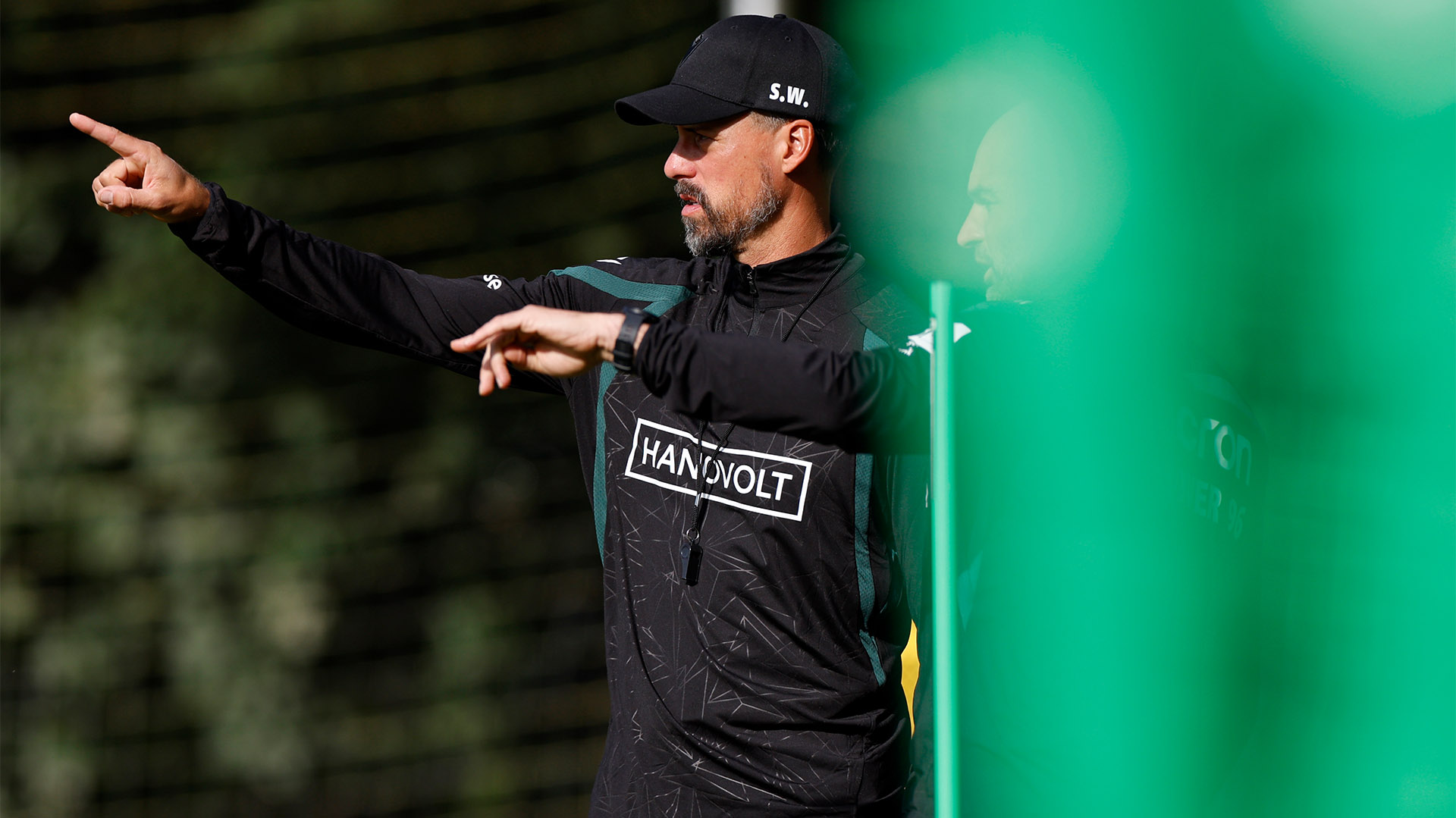 Der 96-Trainer Sandro Wagner zeigt mit dem rechten Zeigefinger, neben ihm steht eine kaum erkennbare Person, die in die gleiche Richtung deutet. 
